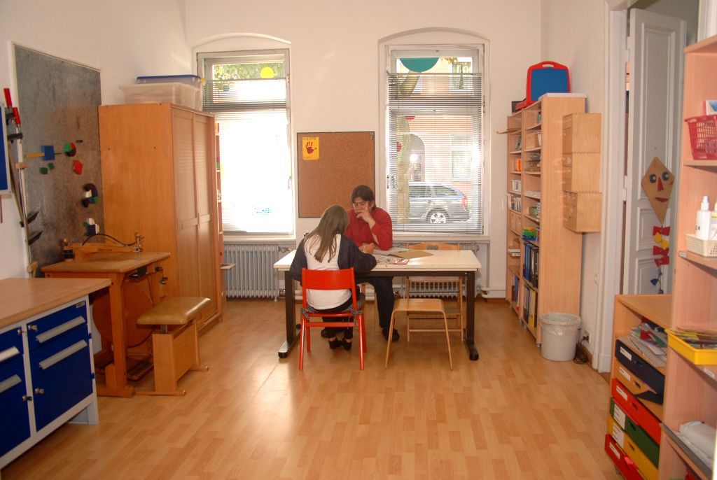 Zwei Personen sitzen an einem Tisch in einem hellen Klassenzimmer mit Holzschränken und bunten Aufbewahrungsboxen.