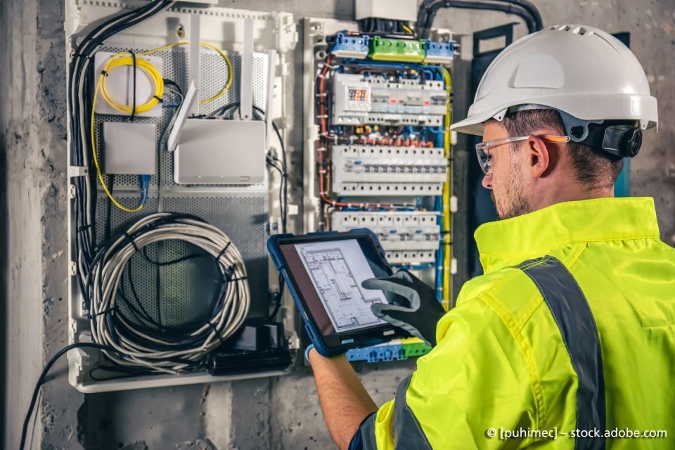 Mann mit Helm vor Verteilerkasten überprüft auf Tablet die Stromversorgung
