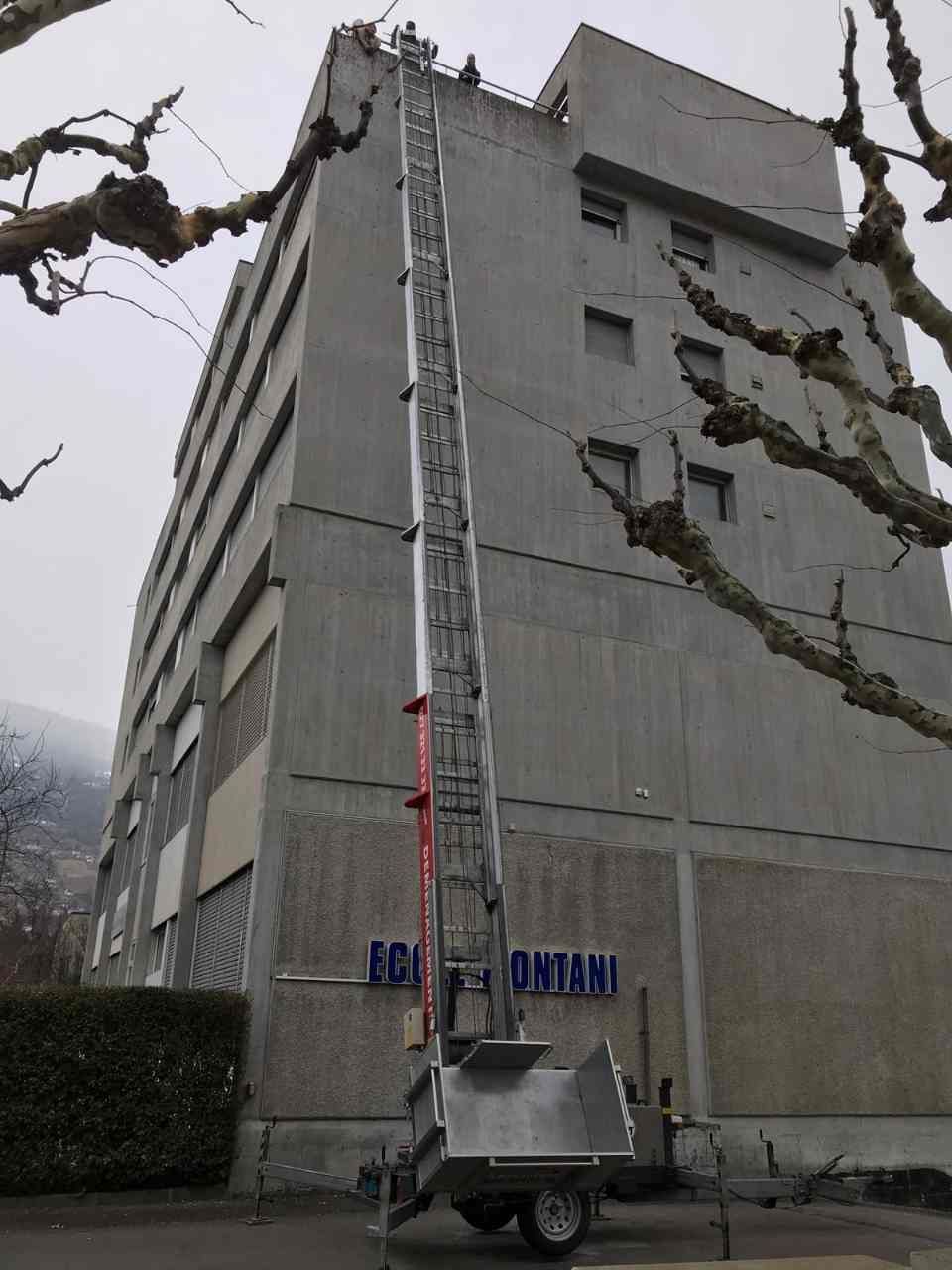 Une remorque avec une échelle attachée utilisant un monte-meuble est garée devant un grand bâtiment