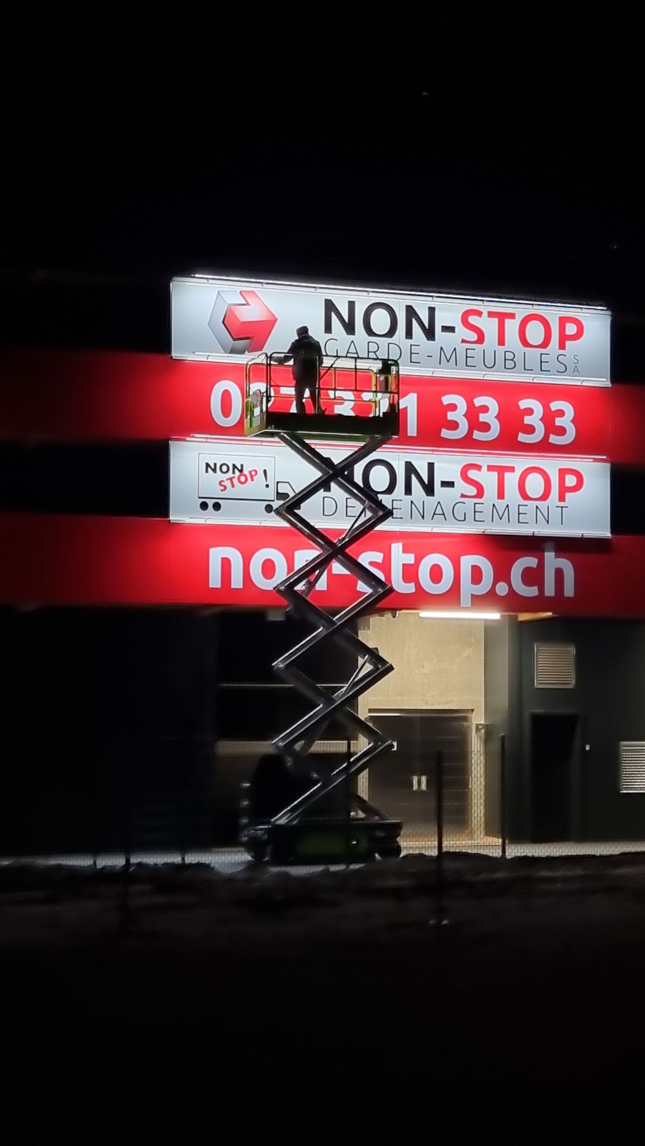 Un homme se tient debout sur une nacelle à ciseaux devant un panneau indiquant non-stop