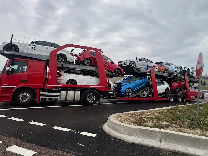 Camión transportador de coches rojos cargado con varios deportivos, circulando por una carretera bajo un cielo nublado