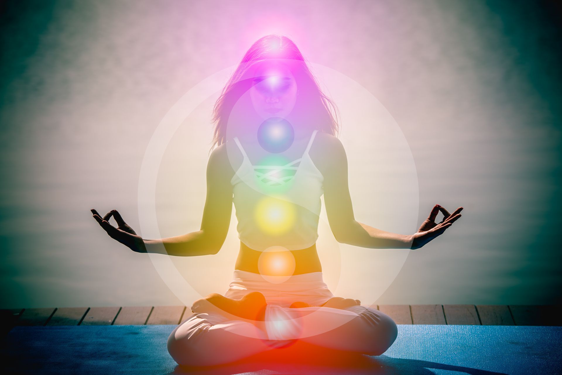 Une femme est assise en position du lotus sur un tapis de yoga avec un arc-en-ciel de chakras qui l'entoure.