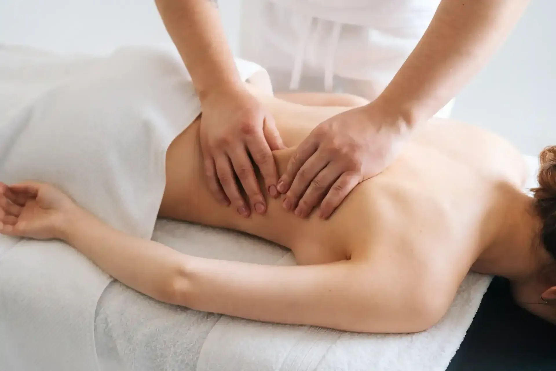 Una persona recibe un masaje de espalda en una camilla. Las manos masajean la espalda, cubierta por una sábana blanca.