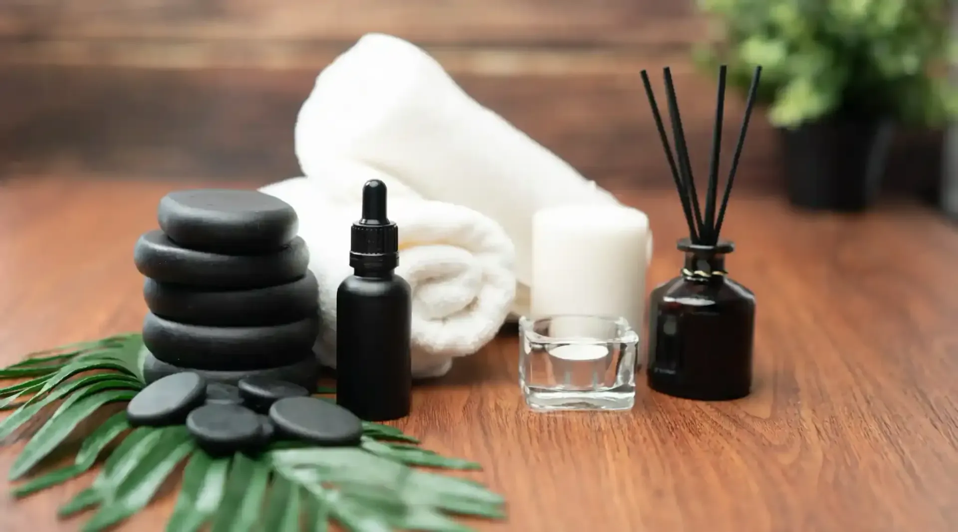 Ambiente de spa con piedras calientes, aceite esencial, toalla, vela y difusor sobre una superficie de madera.