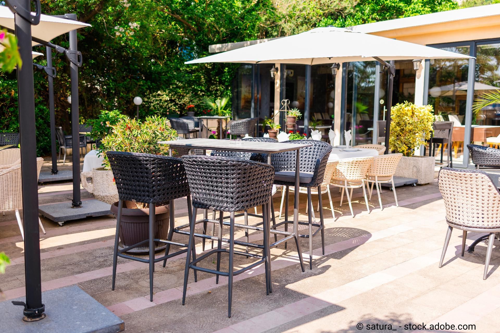 Sonnige Terrasse eines Cafés mit modernen Rattanmöbeln, Sonnenschirmen und viel Grün im Hintergrund