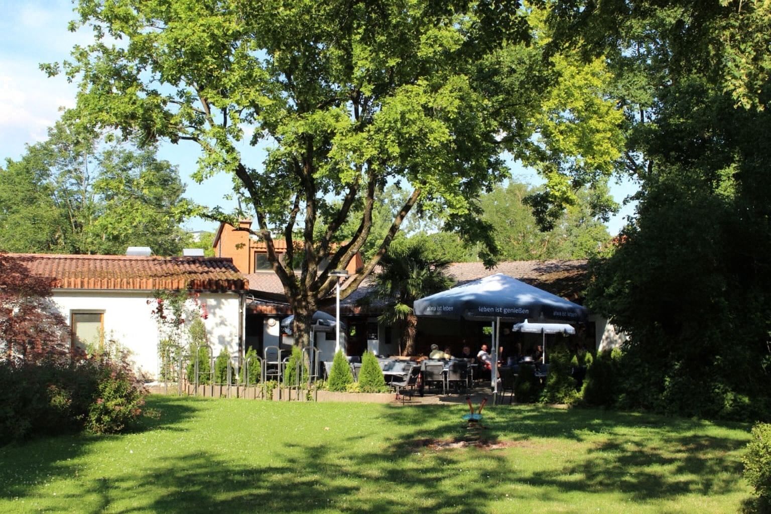 Sonnige Terrasse eines Cafés mit modernen Rattanmöbeln, Sonnenschirmen und viel Grün im Hintergrund