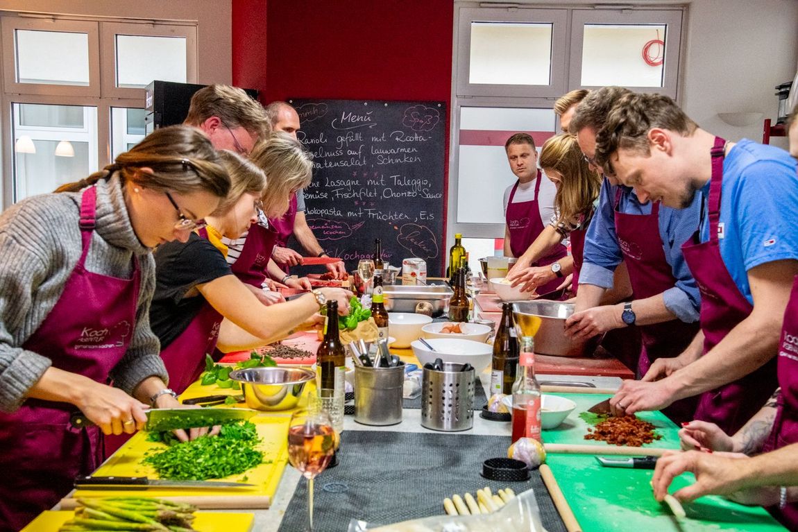 Menschen in Schürzen bereiten bei einem Kochkurs Speisen zu, Gemüse liegt auf Schneidebrettern und an der Tafel steht eine Speisekarte.