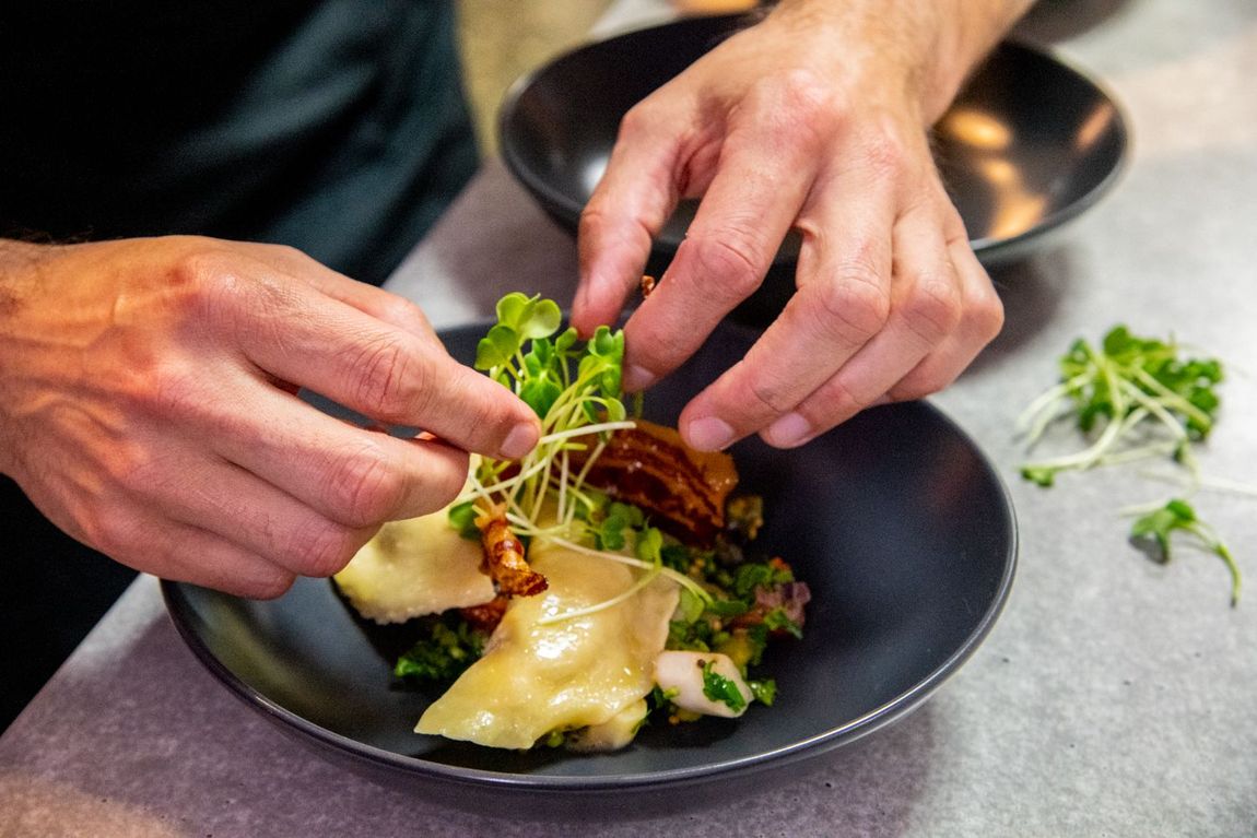 Ein Koch richtet einen Salat an und arrangiert Microgreens. Nahaufnahme von Händen, schwarzer Schüssel und heller Arbeitsfläche.