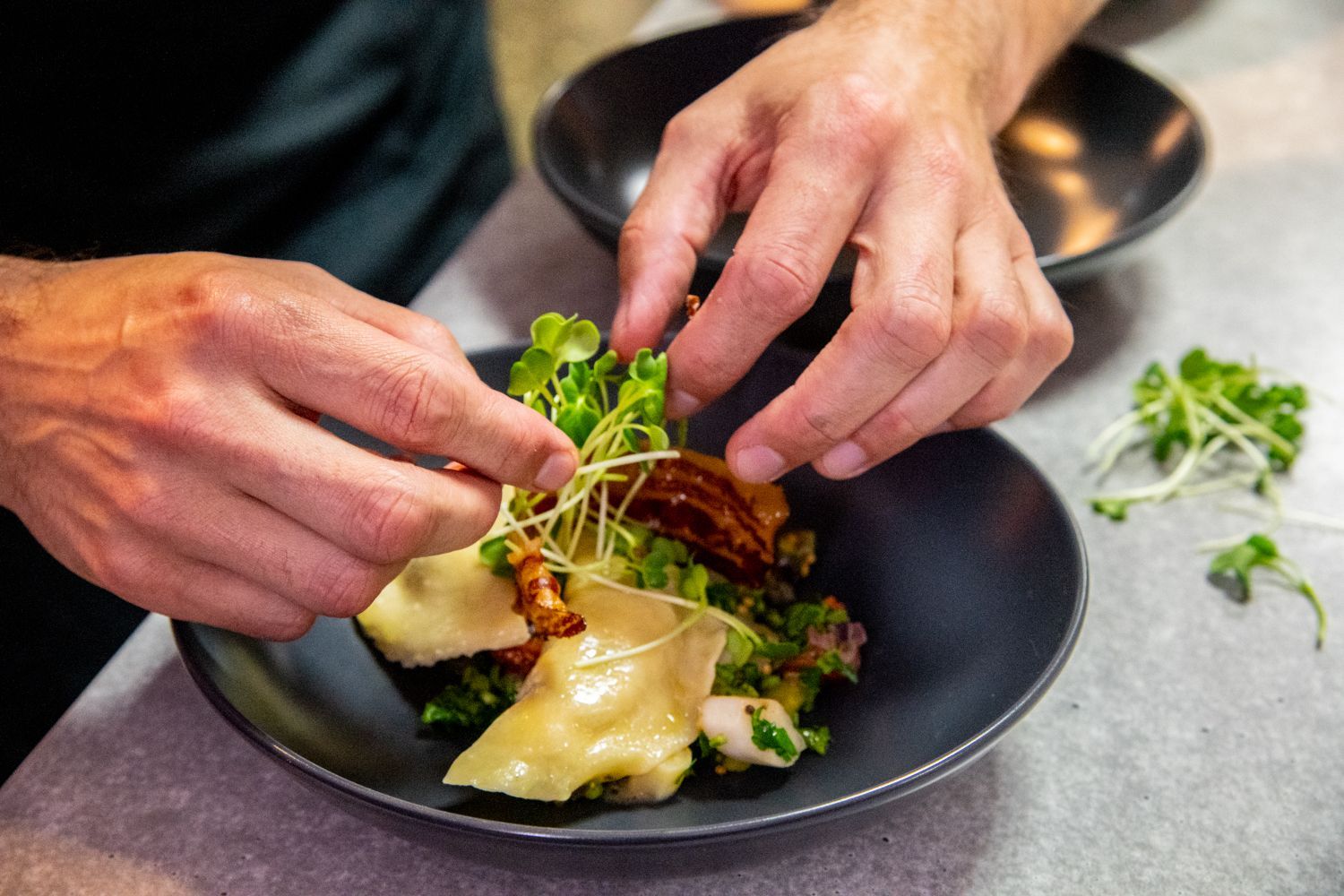 Die Hände des Kochs garnieren einen Salat mit Sprossen in einer dunklen Schüssel auf einer Theke.