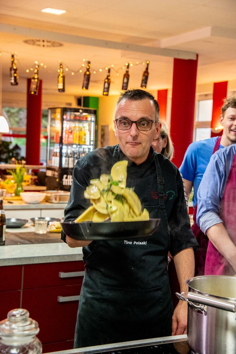 Ein Koch hält einen Topf mit Essen in der Hand und wirkt aufgeregt. Rote Küchenschränke und andere Personen sind zu sehen.