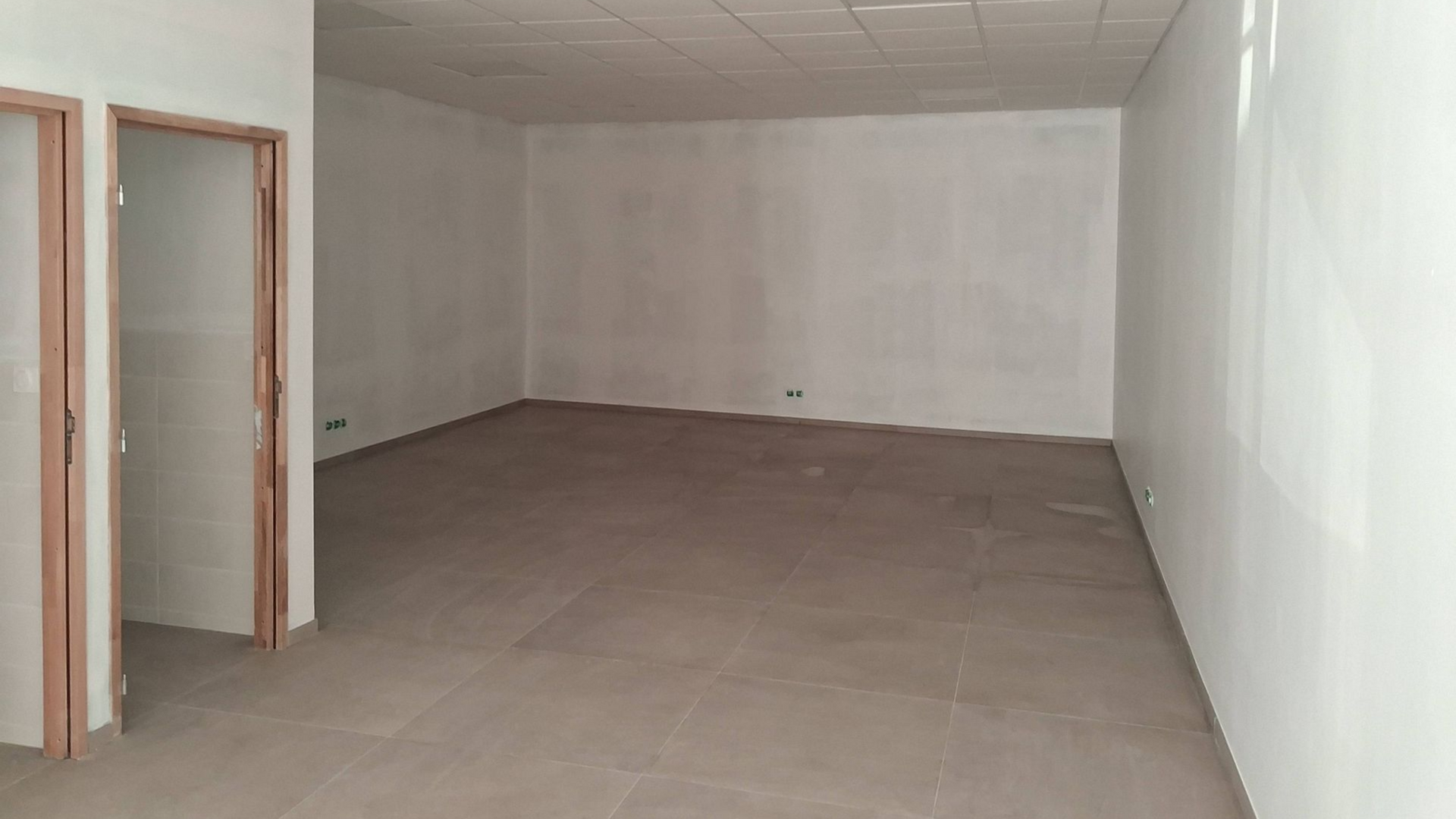 Une pièce vide aux murs blancs unis, au sol carrelé beige, avec un faux plafond et deux encadrements de porte.