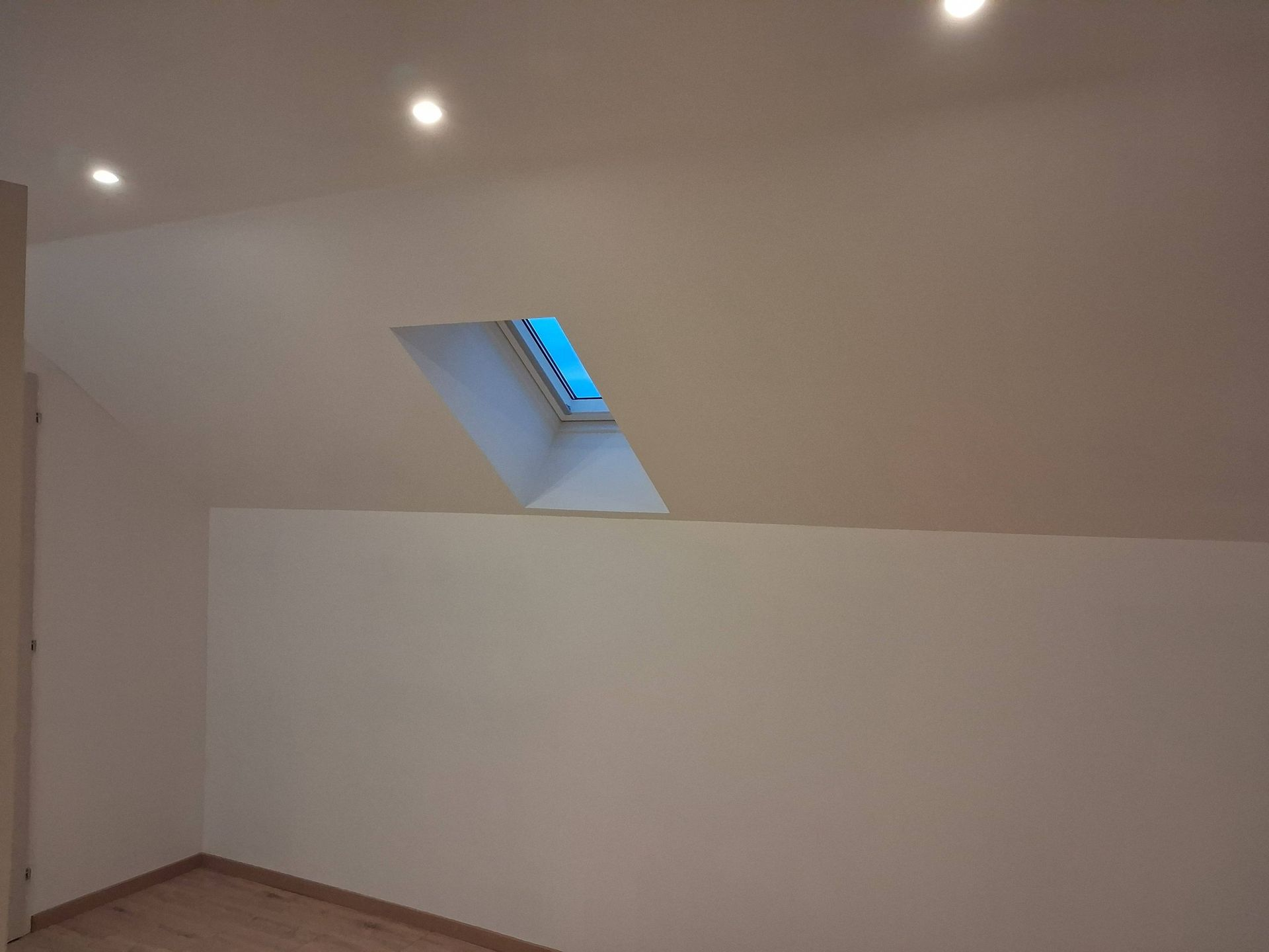 Une pièce mansardée avec des murs beiges, un parquet clair, des spots au plafond et un puits de lumière.