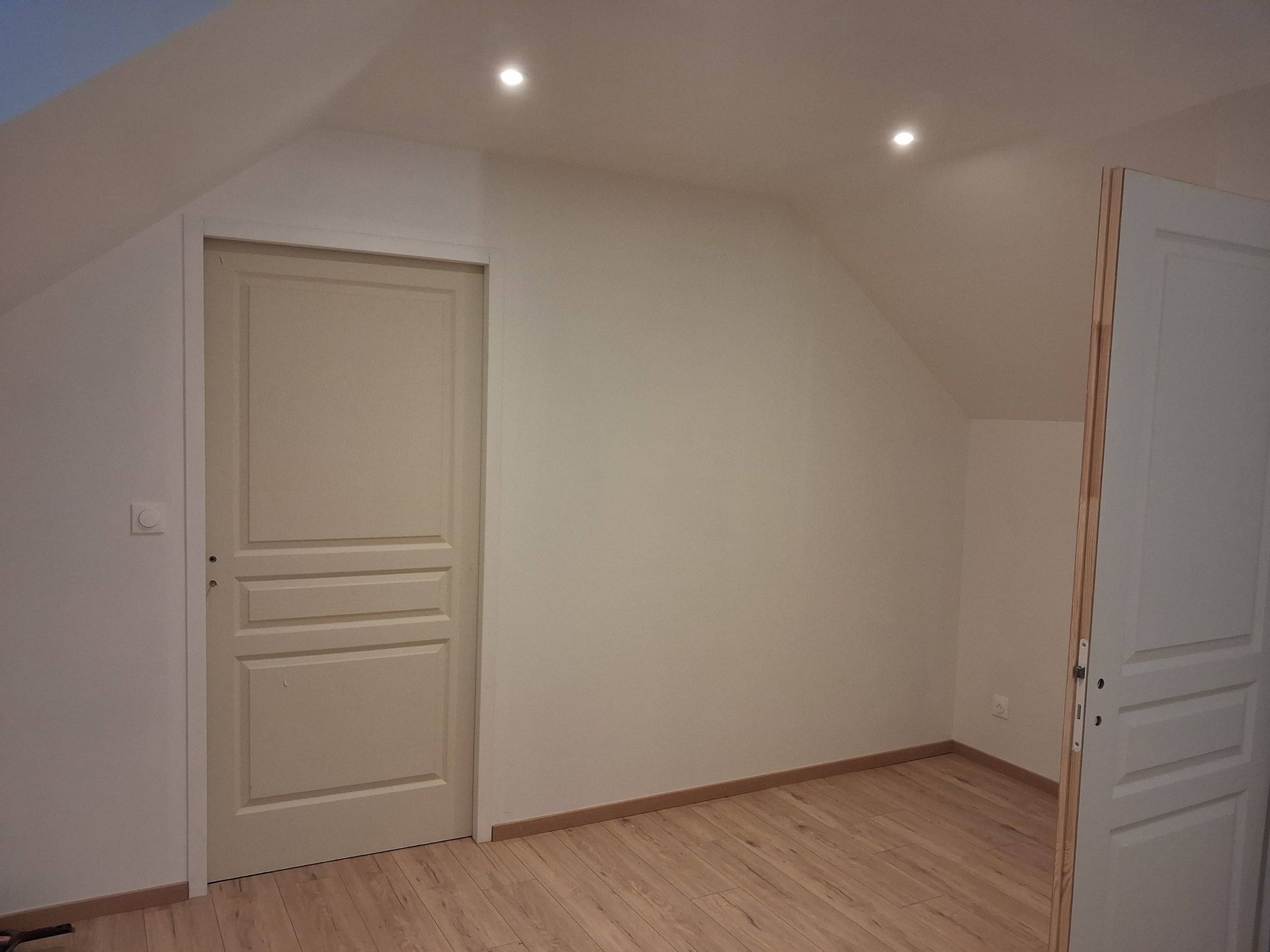Une pièce mansardée avec parquet clair, murs blancs, une porte fermée à gauche et une porte ouverte à droite.