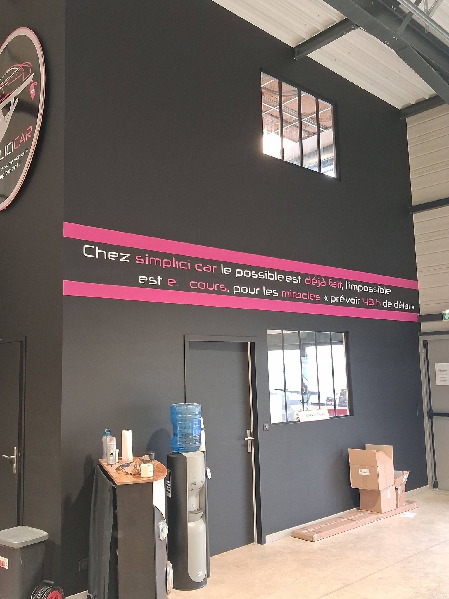 Un grand mur noir dans un espace industriel avec deux fenêtres et des bandes publicitaires roses.