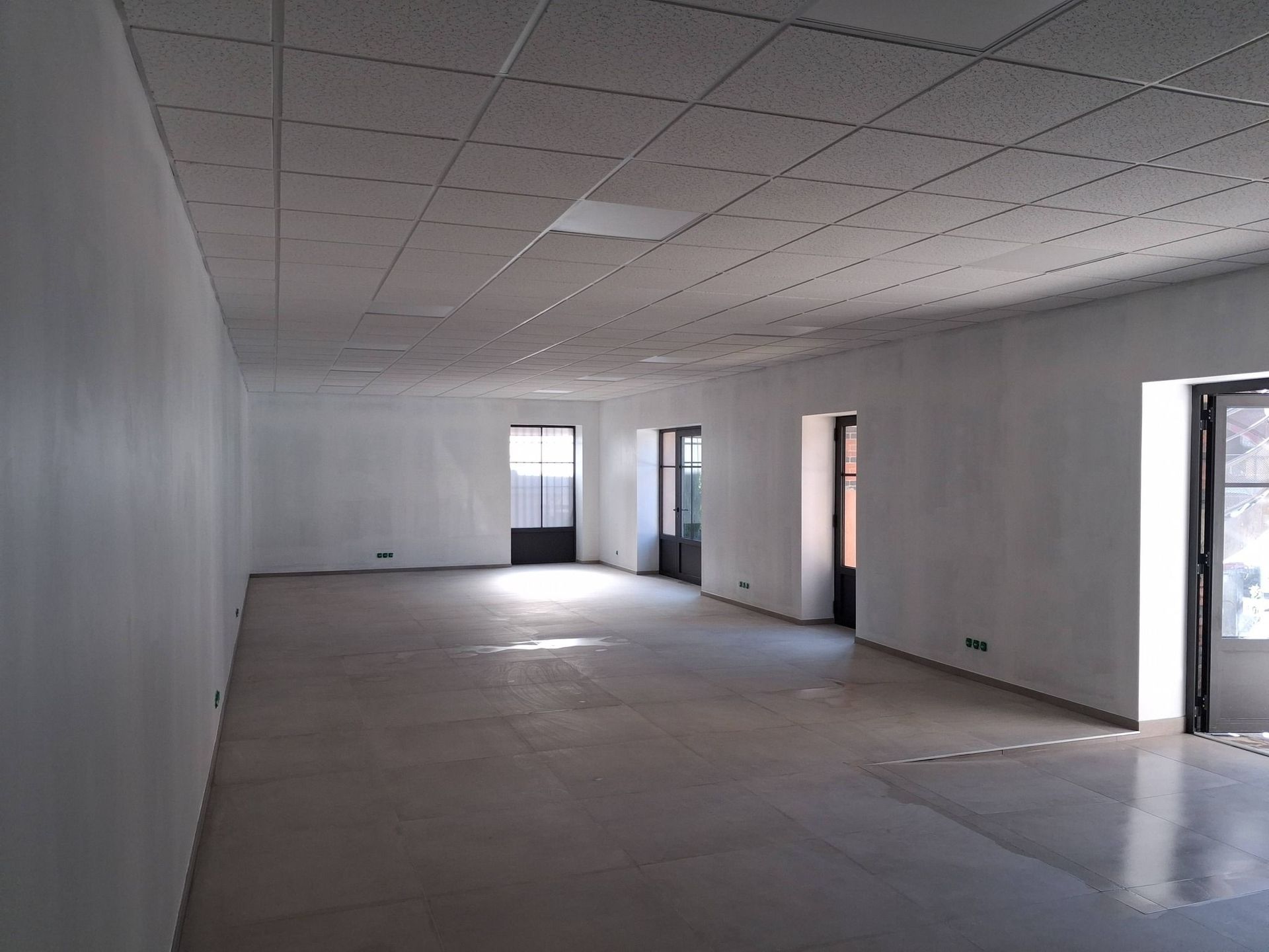 Une pièce minimaliste et vide, aux murs blancs, au sol carrelé gris et au plafond composé de plaques.