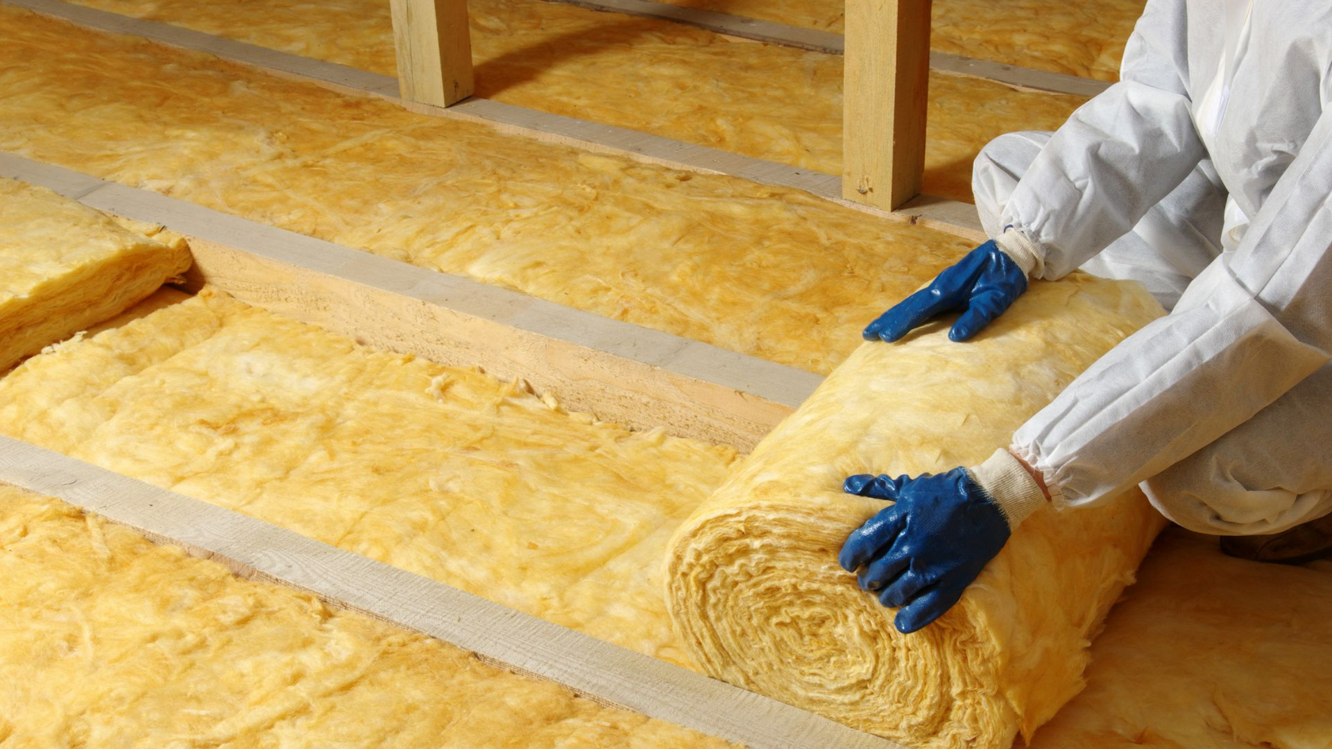 Une personne portant un équipement de protection installe de l'isolant en fibre de verre jaune sous les combles.
