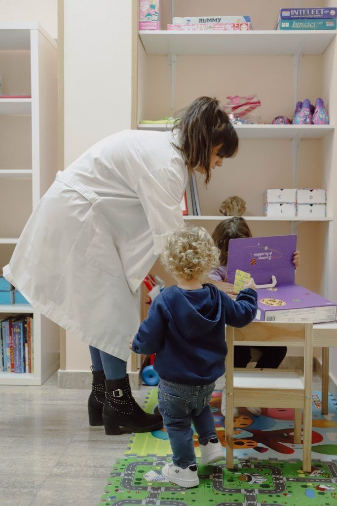Una mujer con bata de laboratorio lee un libro a dos niños en un aula colorida.
