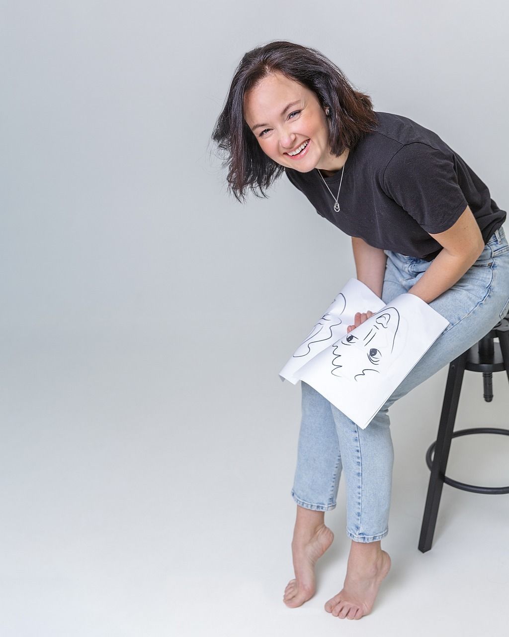 Mujer sonriendo, sosteniendo un dibujo. Está sentada descalza en un taburete, con vaqueros y camiseta negra