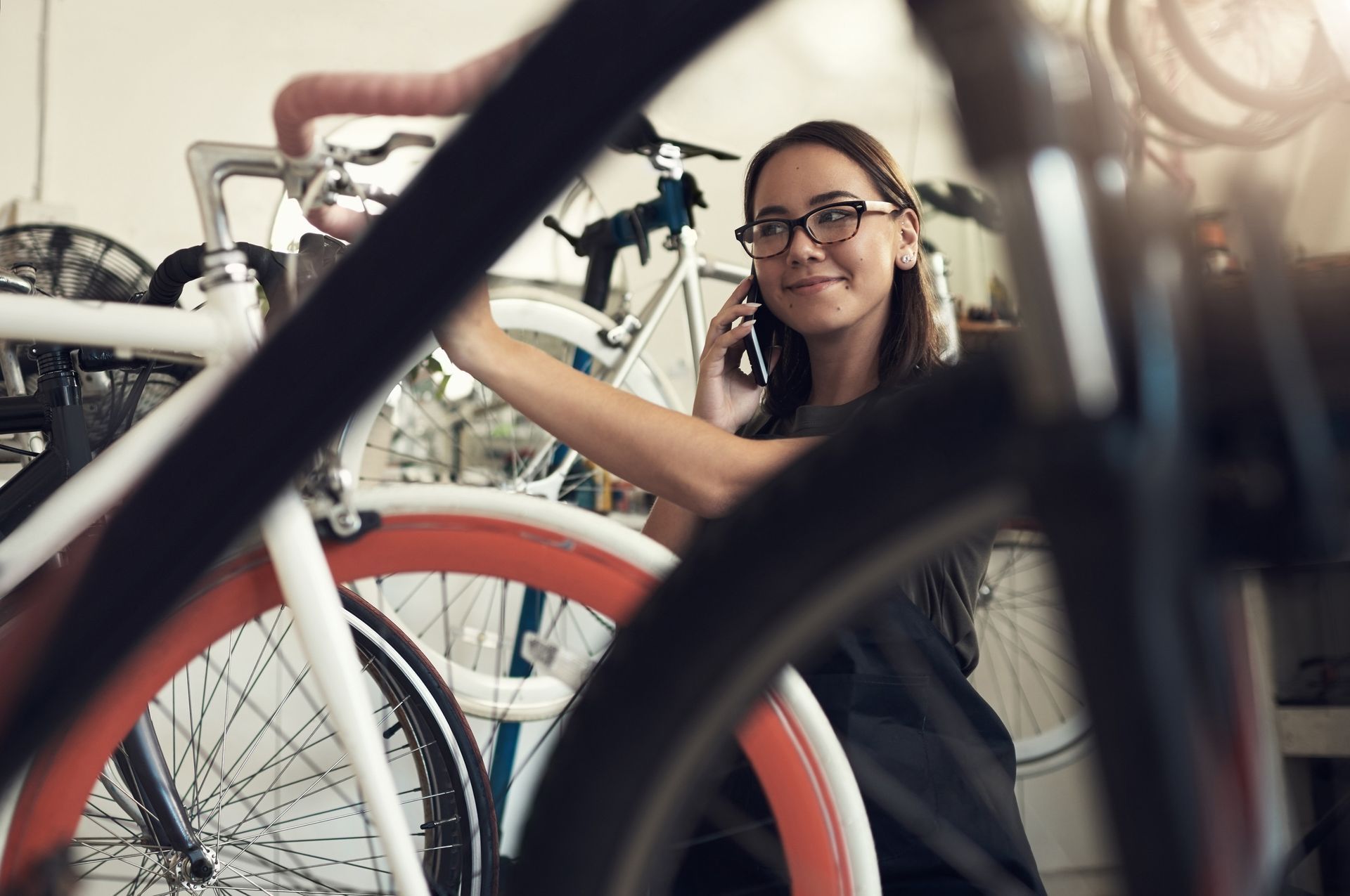 Mujer con gafas en una tienda de bicicletas hablando por teléfono, rodeada de bicicletas.