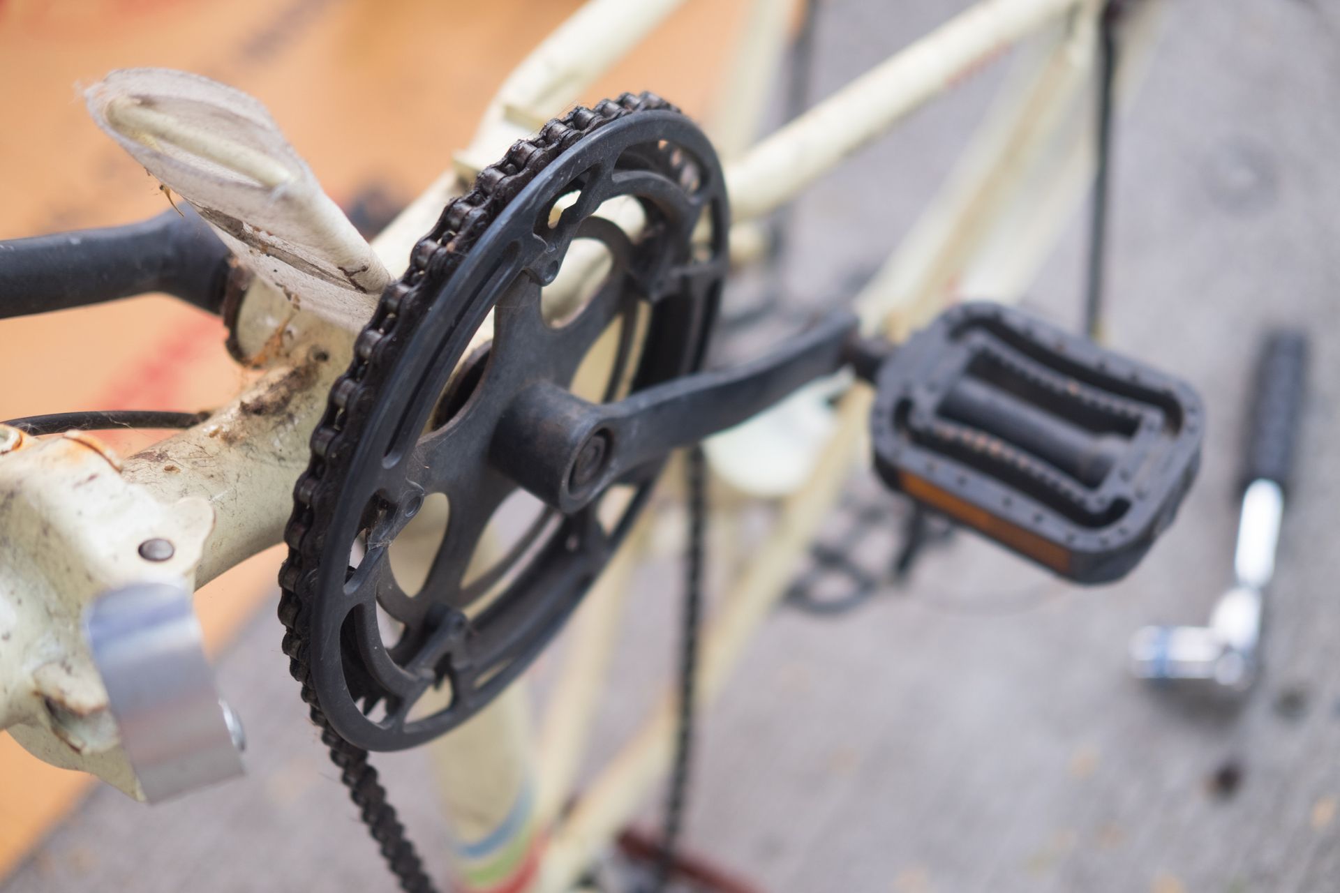 Primer plano de un plato y un pedal de bicicleta de color negro sobre un cuadro de color blanquecino; hay una llave inglesa cerca.