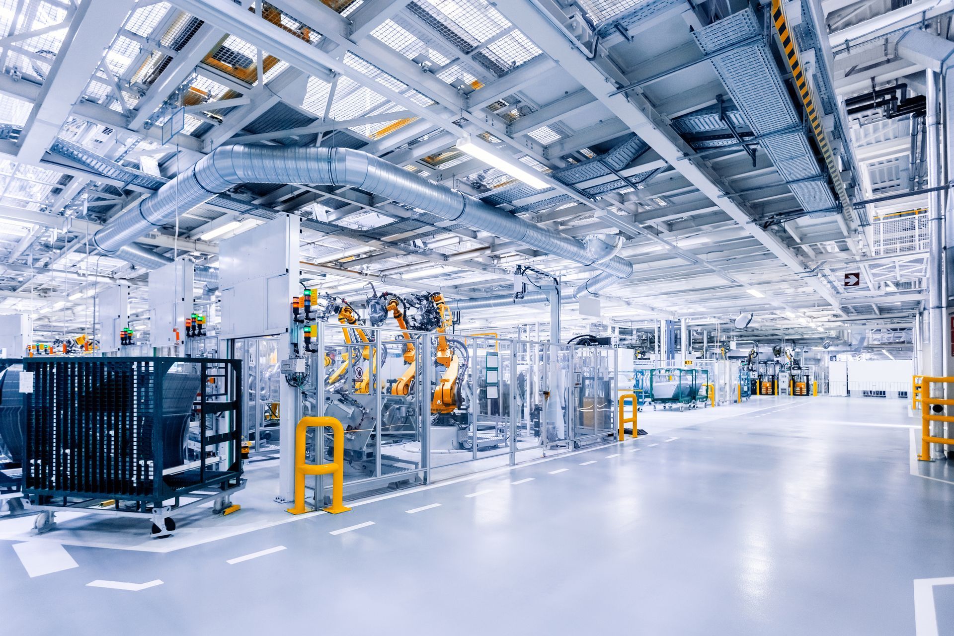 Interno luminoso della fabbrica, macchinari e robot visibili. Pavimento grigio, pareti bianche e soffitto con tubi.