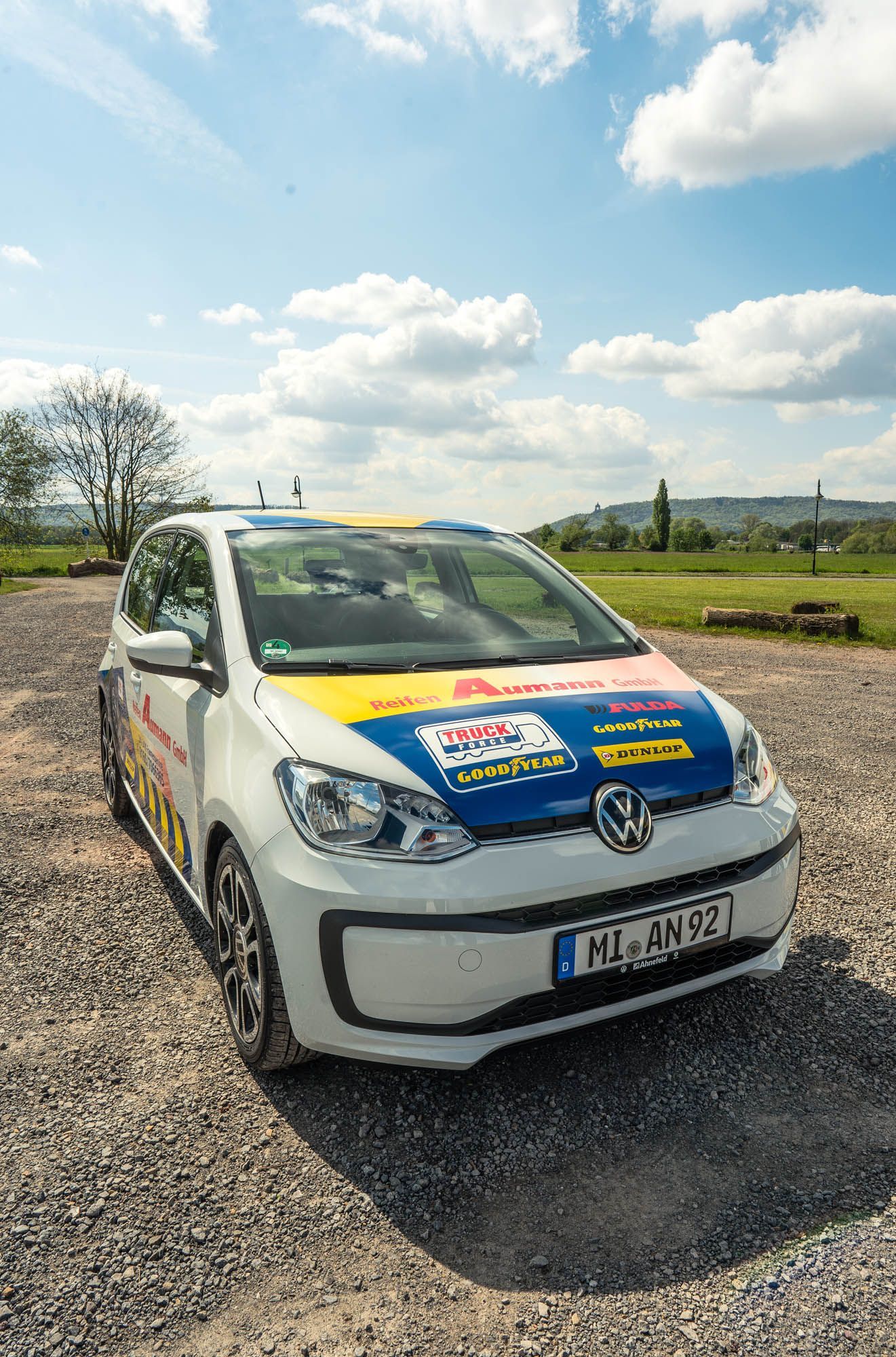 Ein weißes Auto mit blau-gelber Motorhaube steht auf einem Schotterplatz.