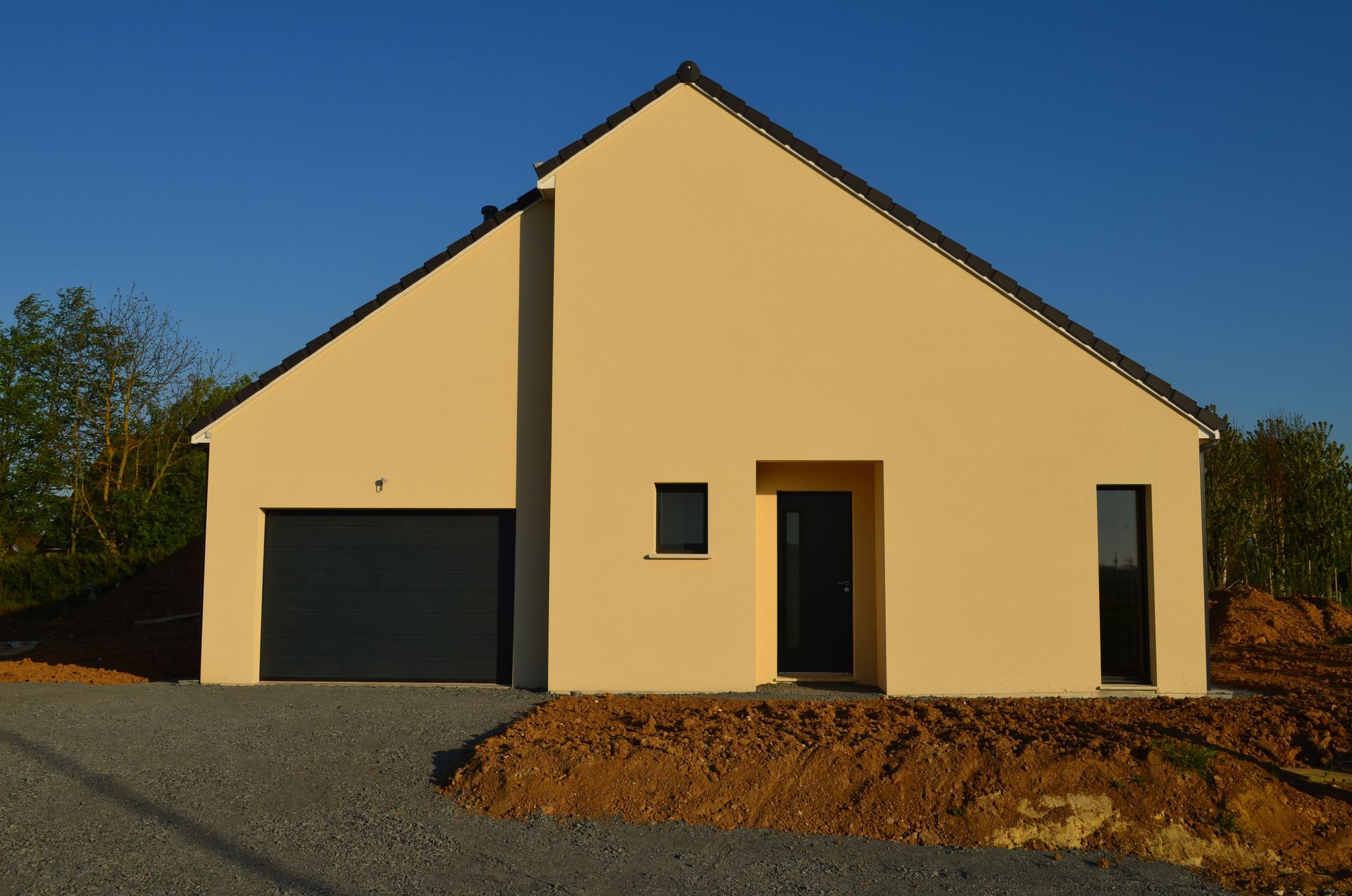 Maison beige avec toit, porte et porte de garage noirs, sur une allée de gravier sous un ciel bleu.