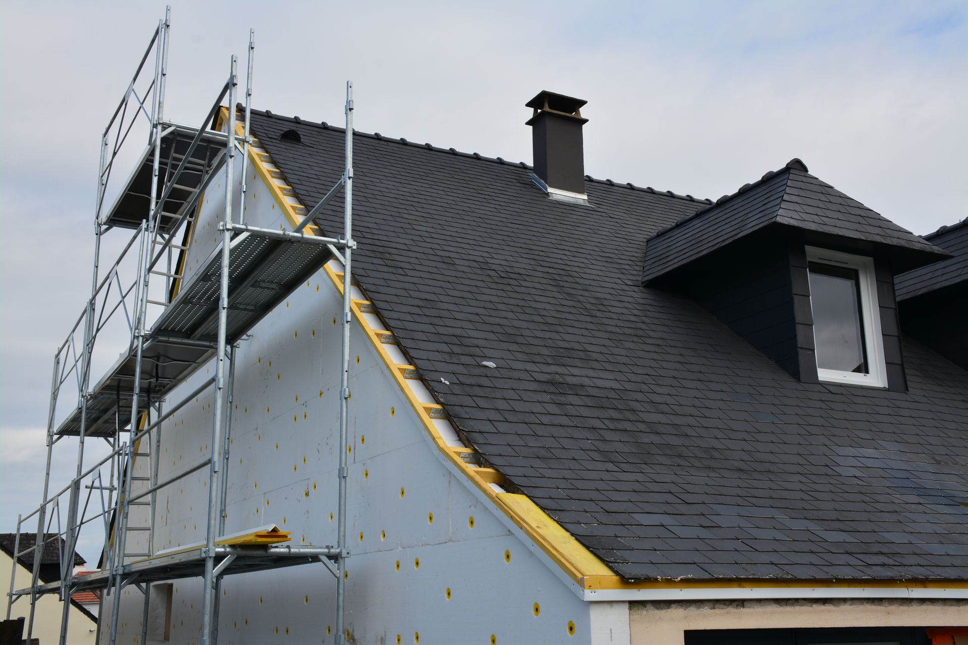 Maison avec échafaudages, mur latéral en construction, toit sombre avec cheminée et lucarne.
