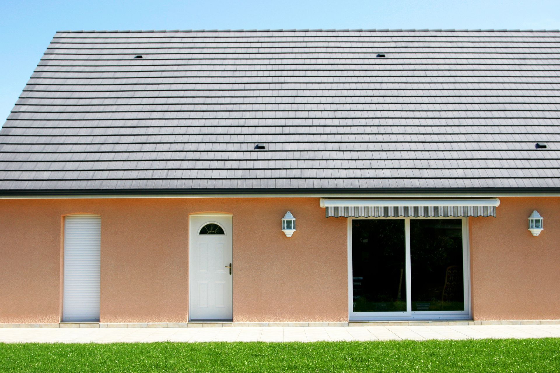 Maison beige avec toit gris, porte et encadrements blancs, et grande porte coulissante en verre avec auvent.