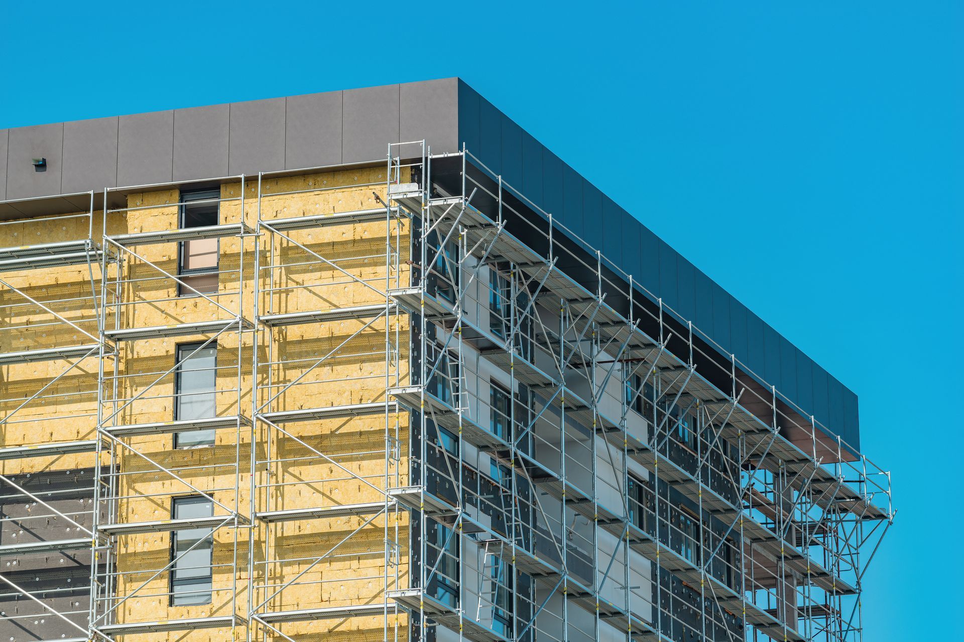 Façade du bâtiment ; installation d’isolants et de panneaux. Échafaudages, fenêtres et matériaux bruns et beiges.