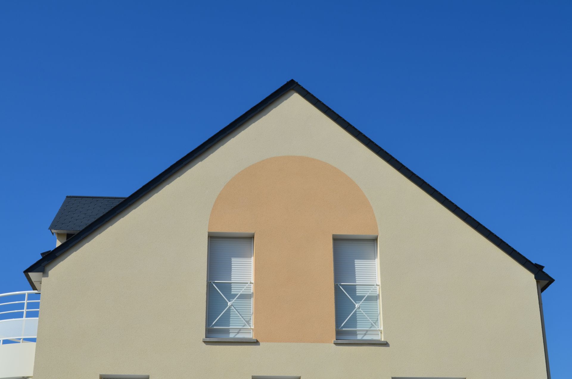 Façade de maison beige avec un toit sombre, deux fenêtres et une structure en arc, sur fond de ciel bleu.