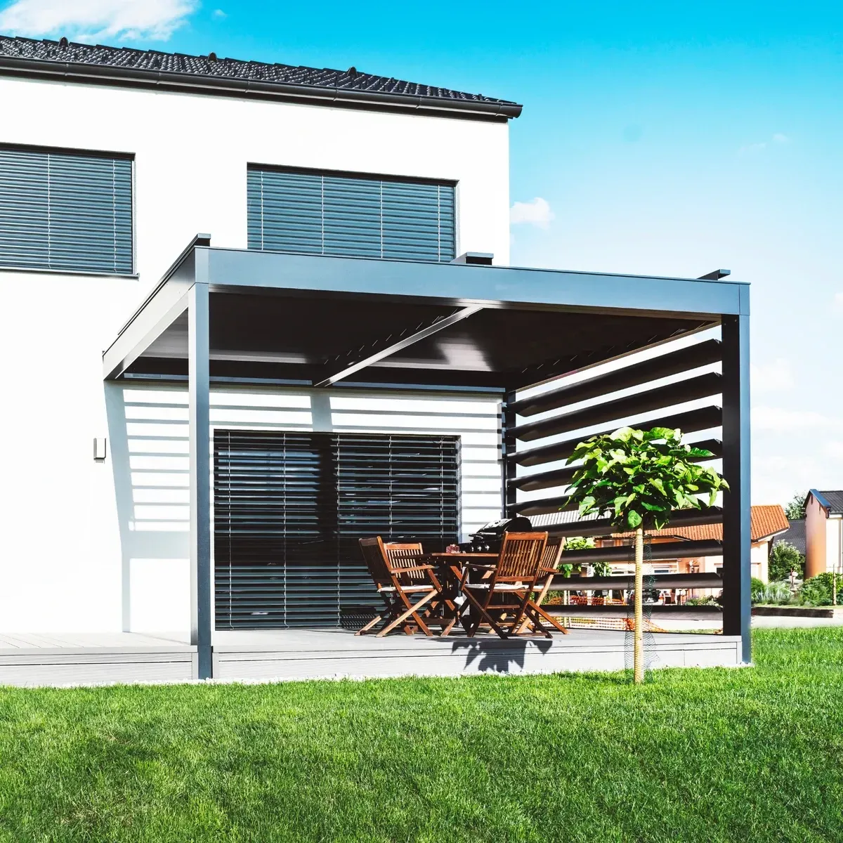 Pergola moderne avec ensemble de salle à manger sur une terrasse à côté d'une maison blanche, par une journée ensoleillée.