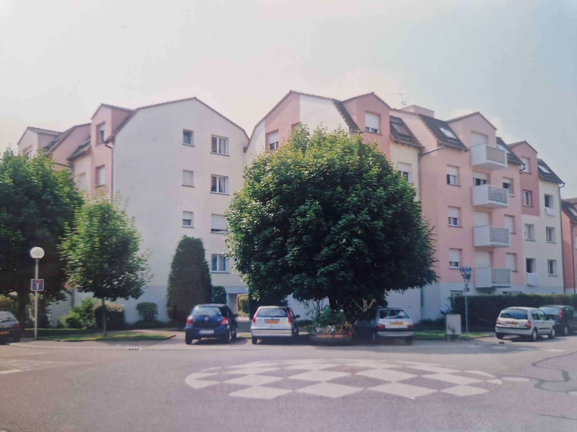 Un complexe d'appartements résidentiels à plusieurs étages avec un grand arbre central, des voitures garées et un motif de route peint à l'avant.