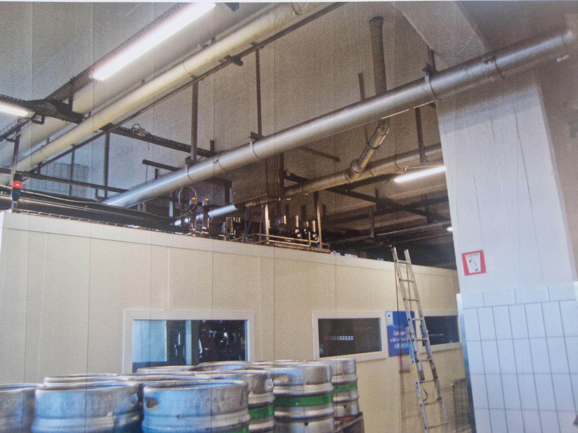 Espace industriel avec des fûts argentés au premier plan, des tuyaux au plafond et une échelle appuyée contre un mur blanc.