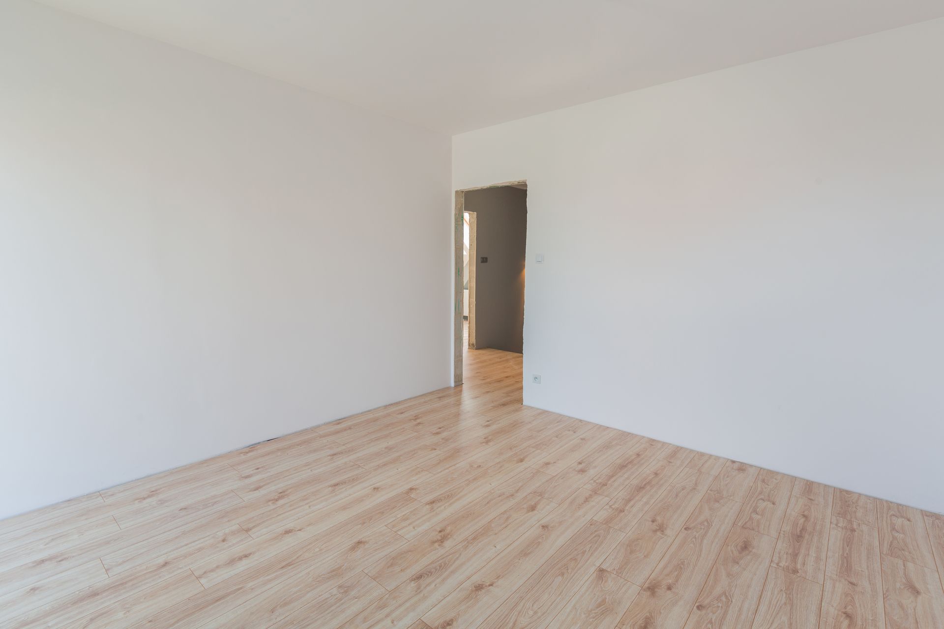 Une pièce vide et minimaliste, aux murs blancs unis et au sol clair à motifs de bois, avec une porte.