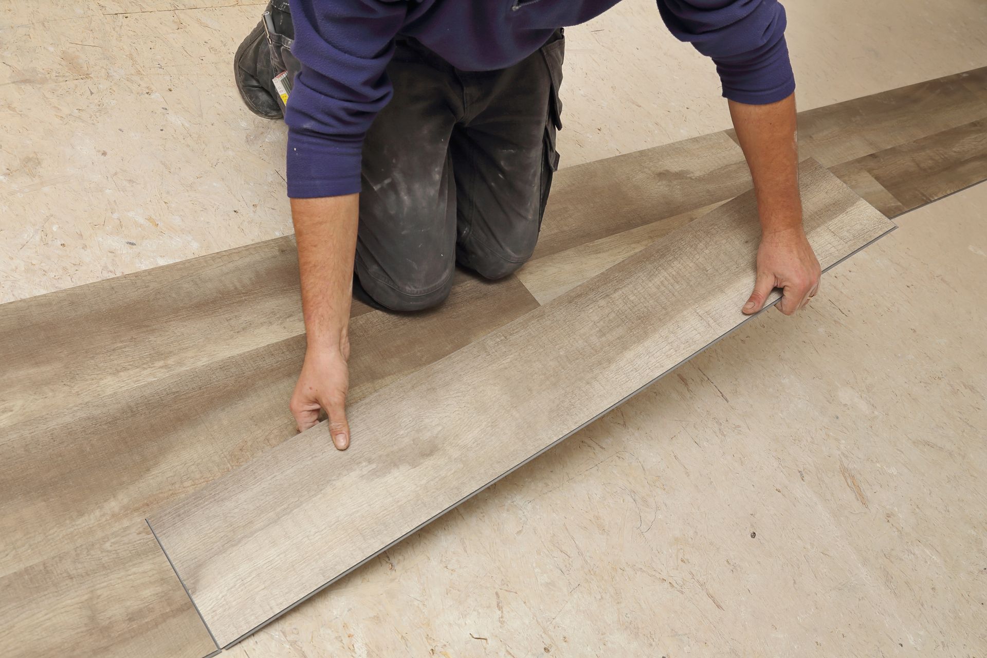 Une personne agenouillée sur un sol en béton installe de longues lames de vinyle imitation bois.