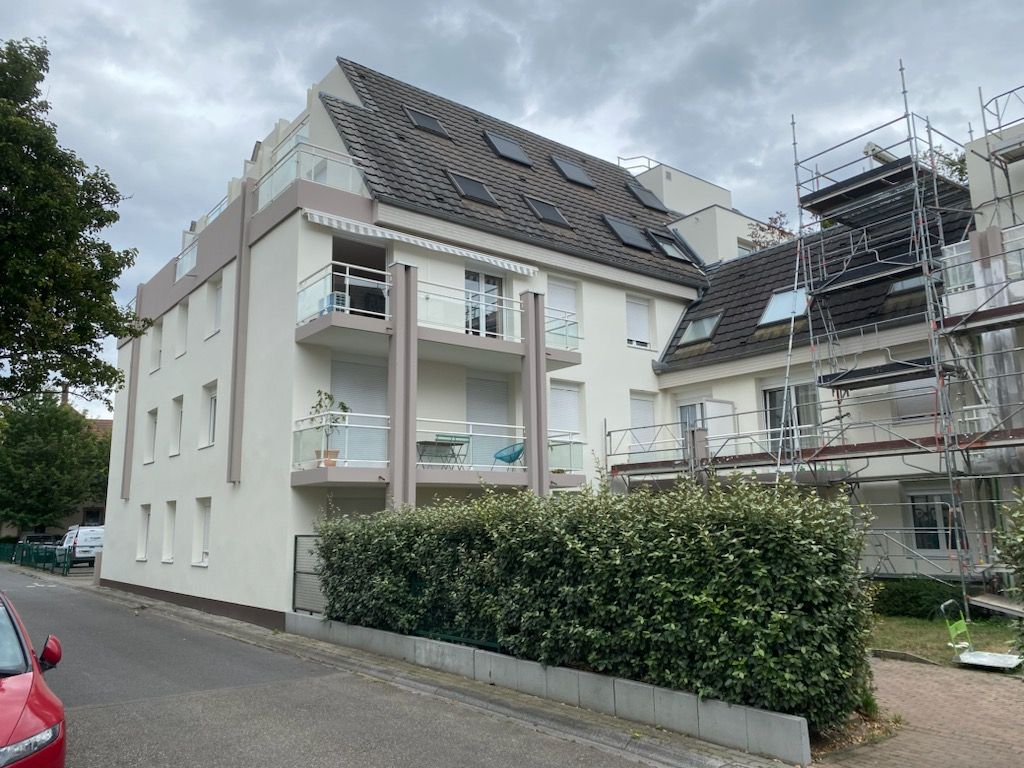 Un immeuble d'appartements blanc à plusieurs étages, avec un toit mansardé en tuiles sombres, des balcons et des échafaudages sur le côté droit.