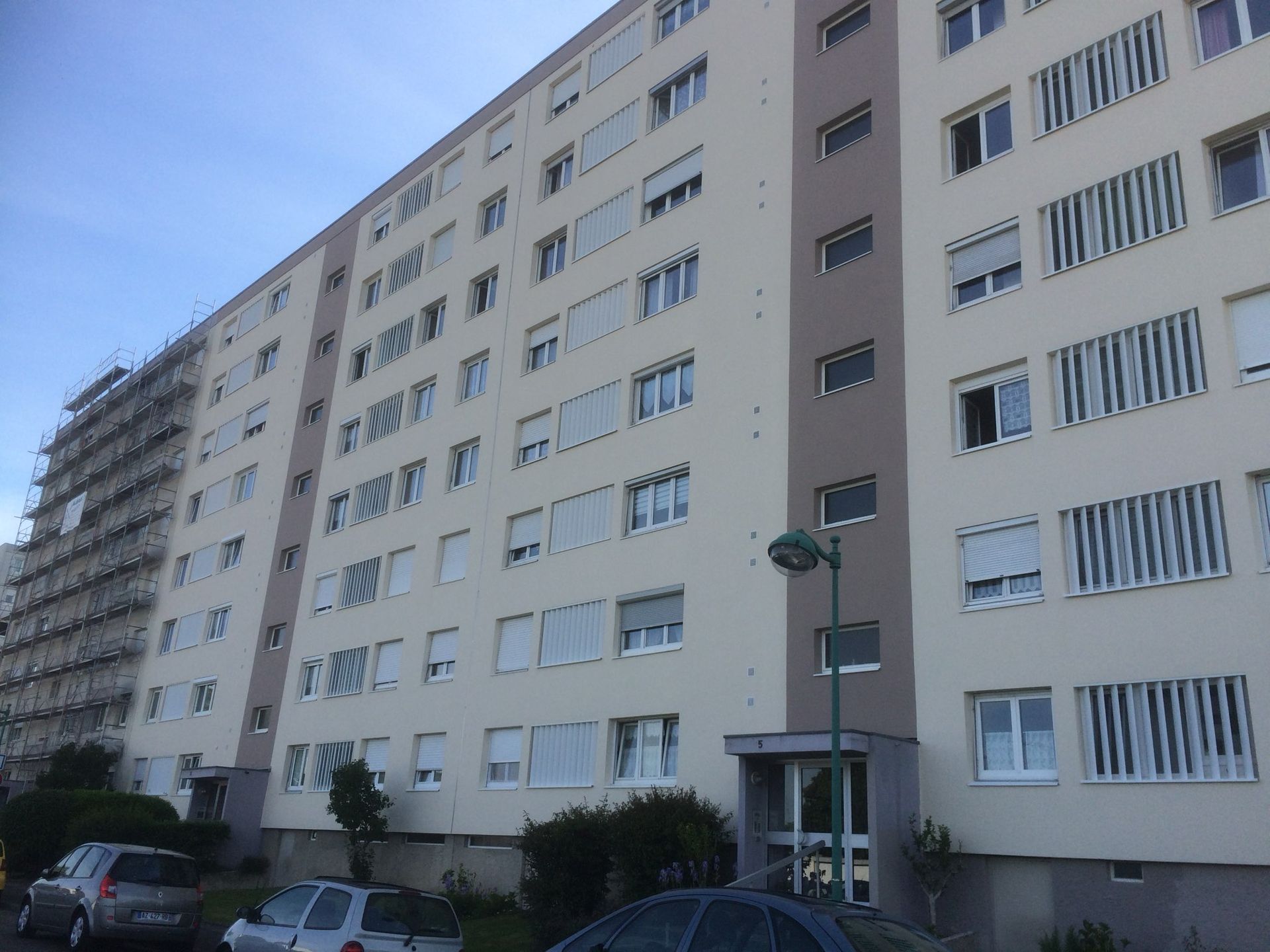 Un immeuble d'appartements à plusieurs étages avec une façade beige, des volets blancs et une structure d'échafaudage métallique sur la gauche.