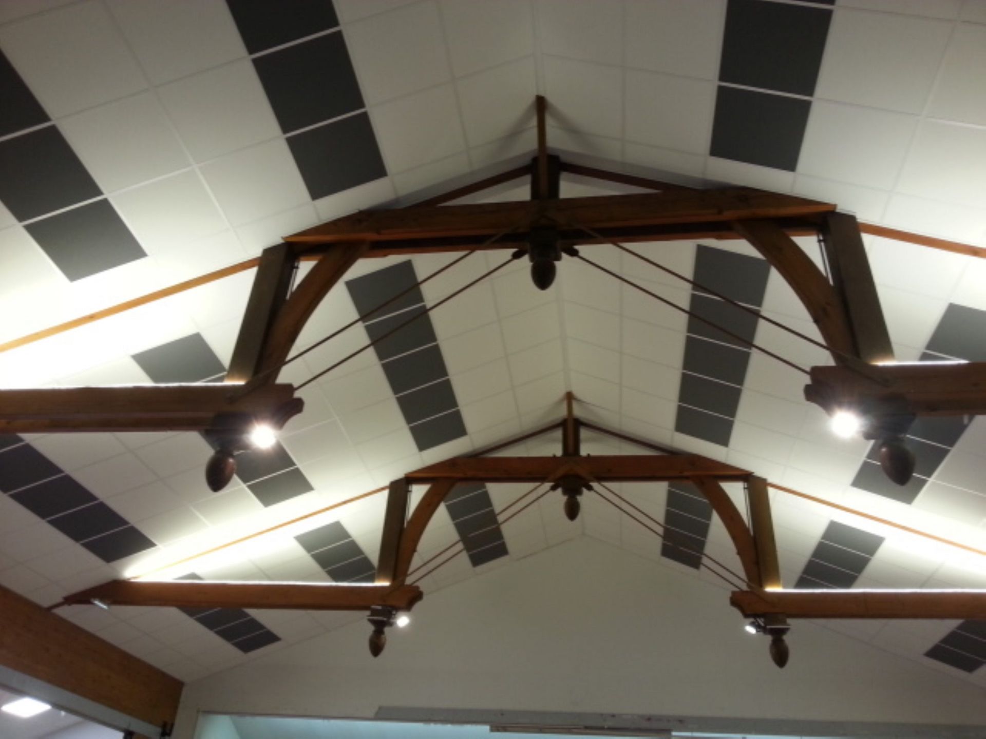 Poutres en bois soutenant un plafond blanc avec des panneaux rectangulaires gris et un éclairage encastré.