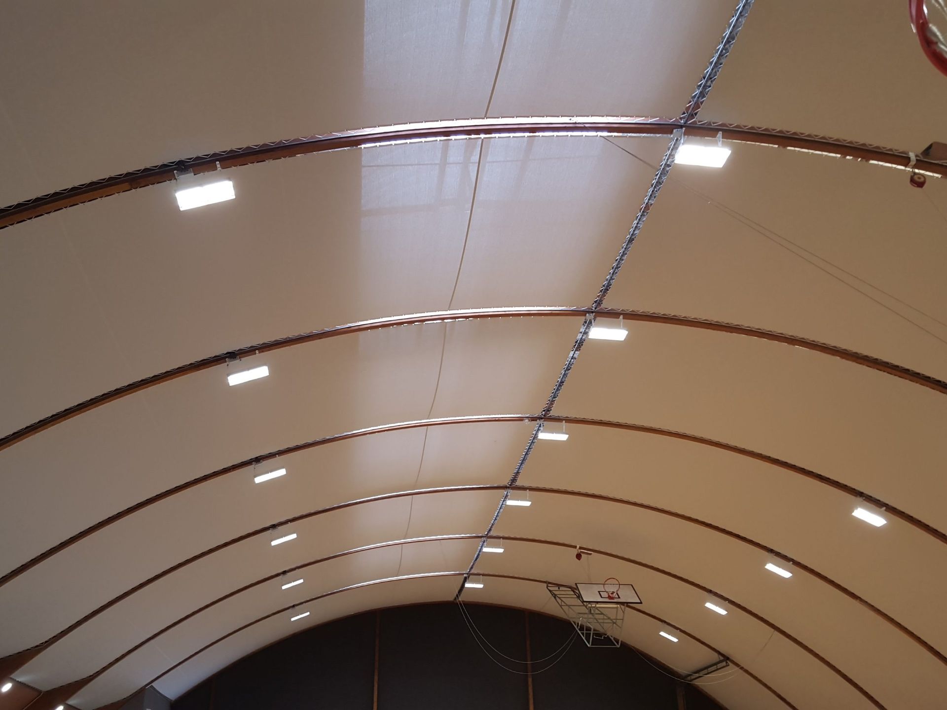 Plafond de gymnase voûté avec lumières rectangulaires, poutres brunes et un panier de basket visible.