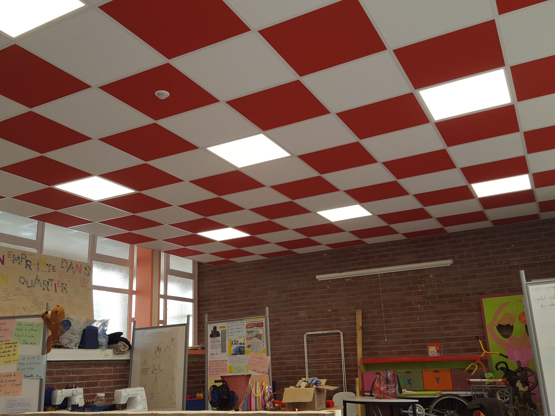 Plafond à carreaux rouges et blancs dans une salle de classe avec fenêtres, mur de briques et divers objets.