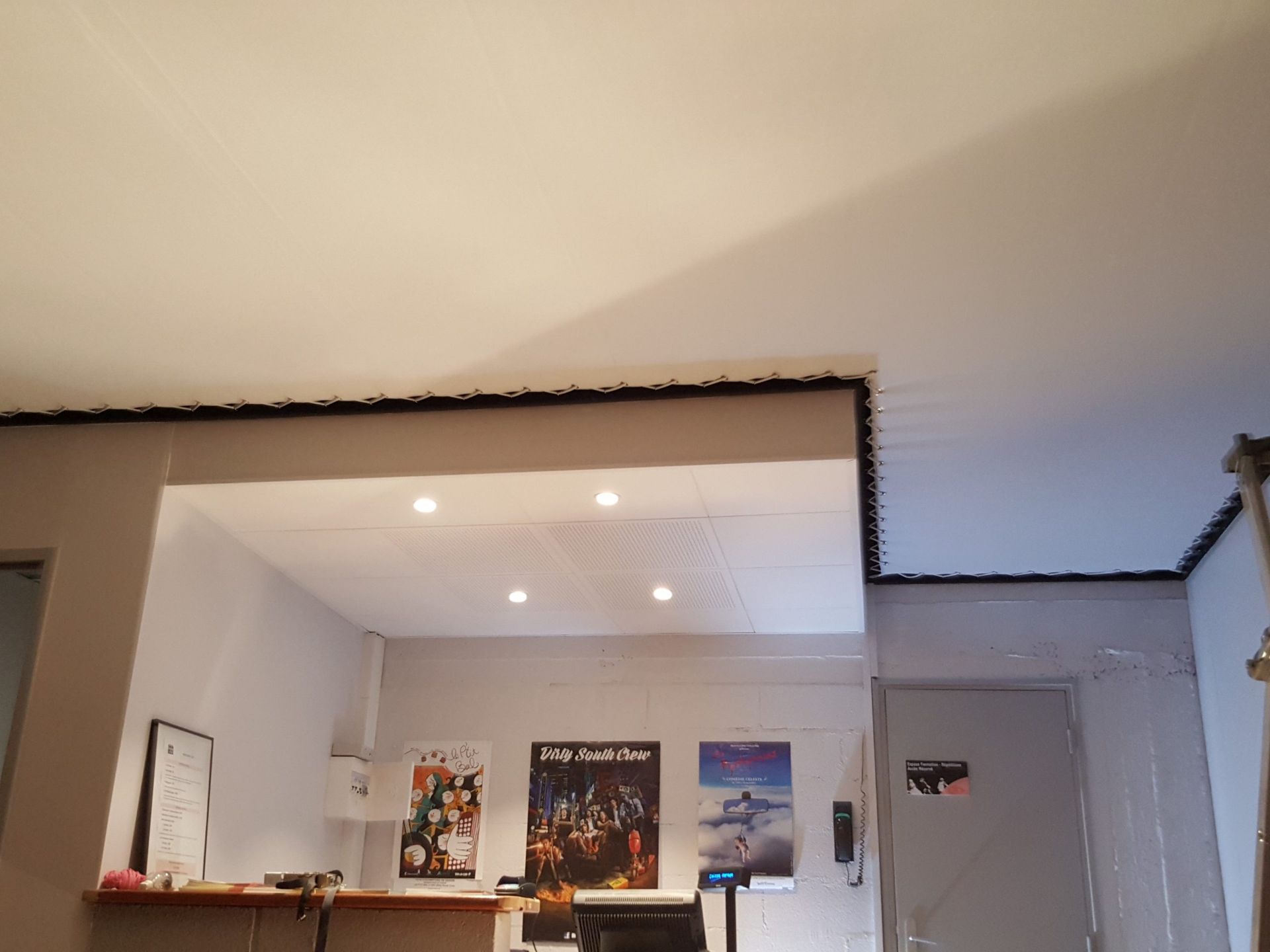 Intérieur avec murs blancs, plafonniers et affiches. Un comptoir et une porte sont visibles.
