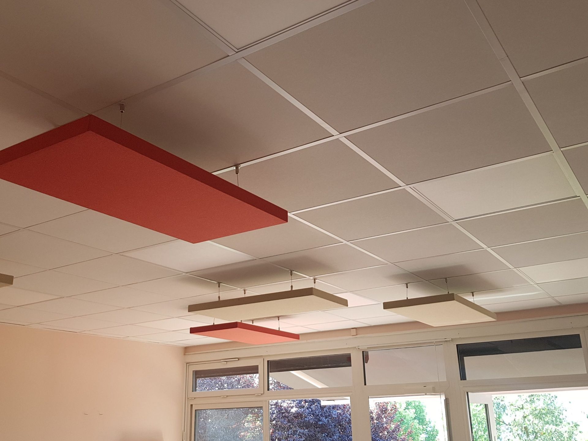 Panneaux acoustiques rouges et beiges suspendus à un plafond blanc à motif quadrillé, près des fenêtres.
