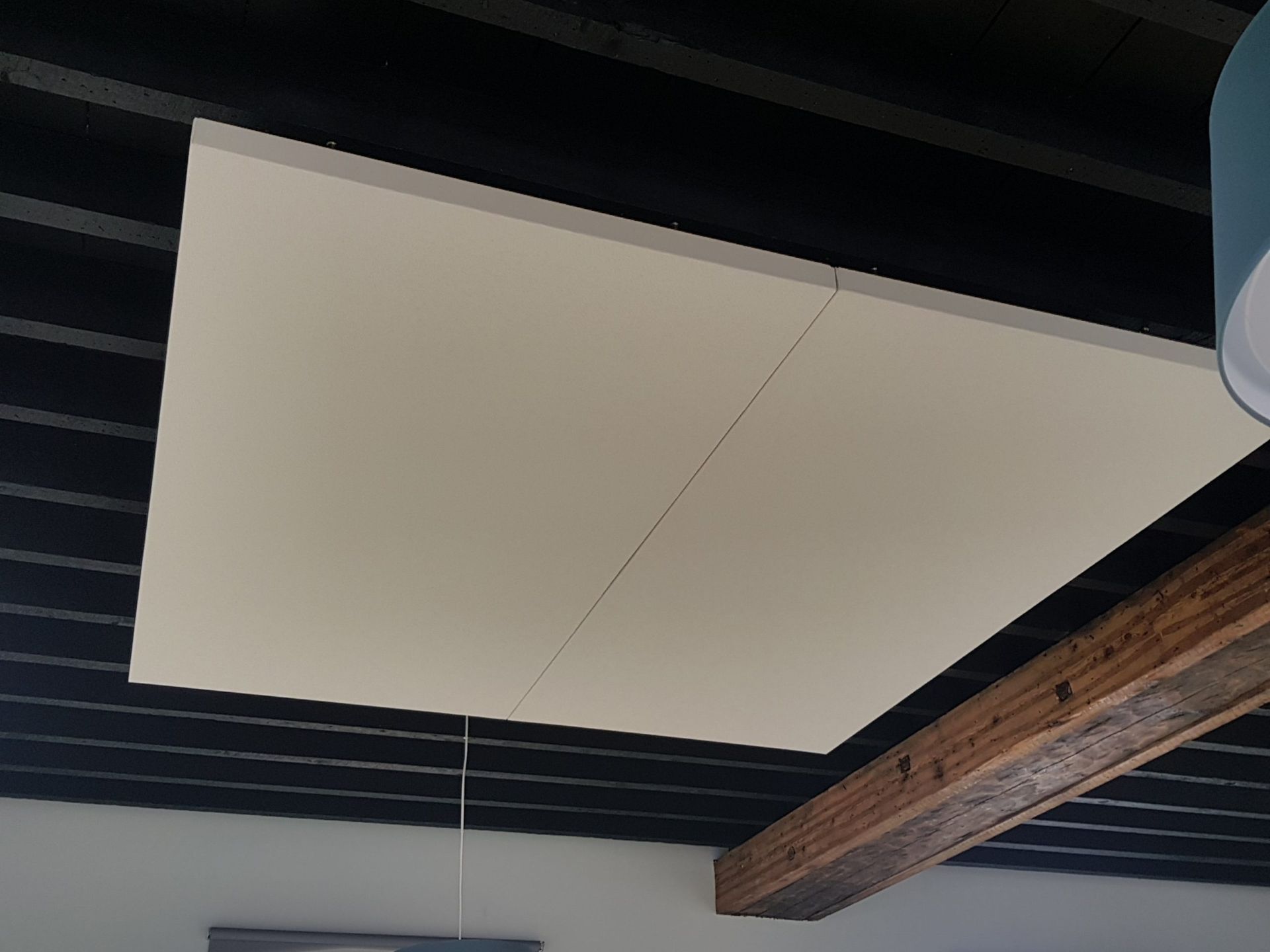 Panneau acoustique couleur crème fixé à un plafond noir. Des poutres en bois sont visibles en dessous.