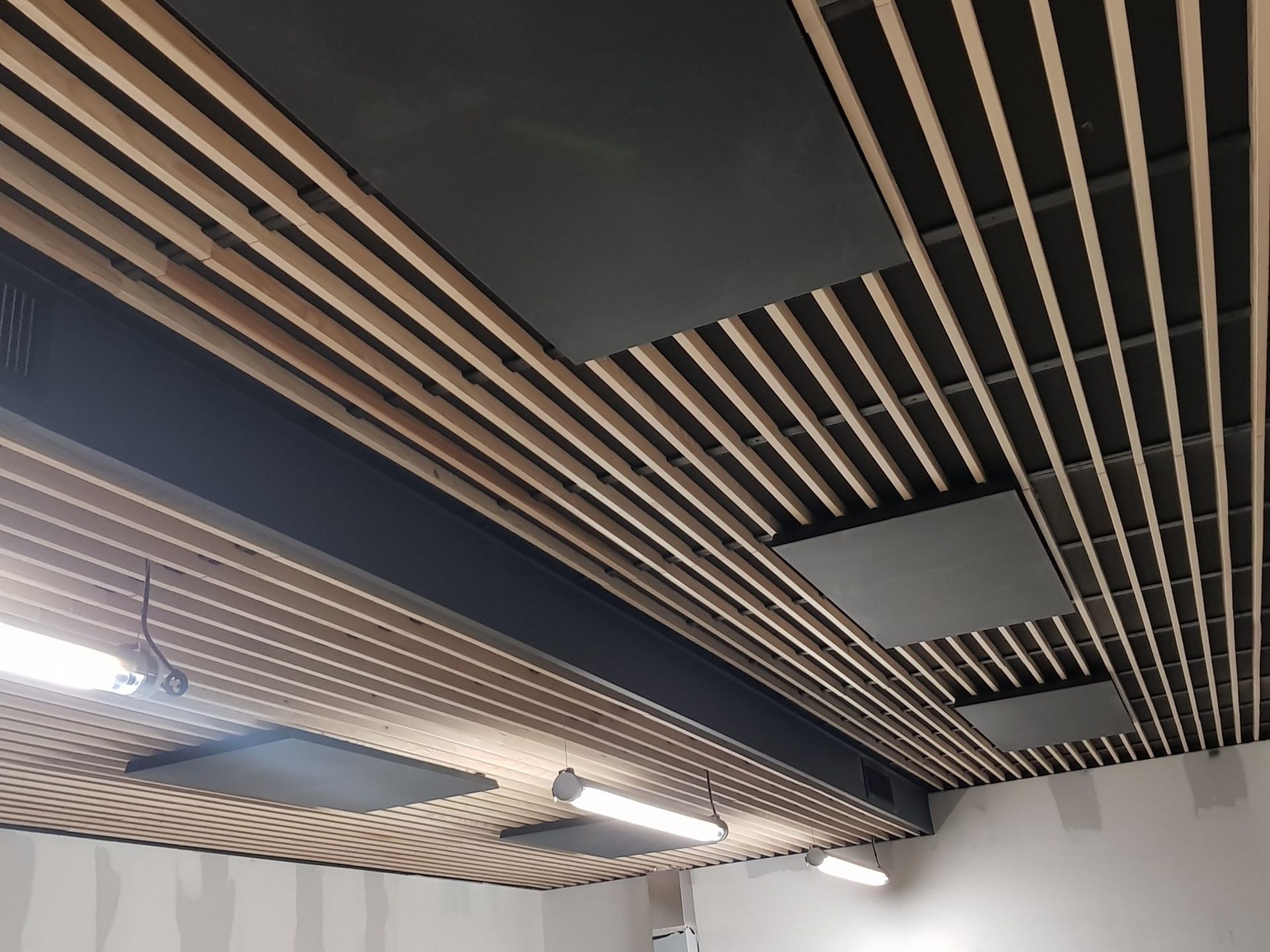 Plafond à lattes de bois avec panneaux insonorisants gris foncé et lumières fluorescentes dans un intérieur moderne.