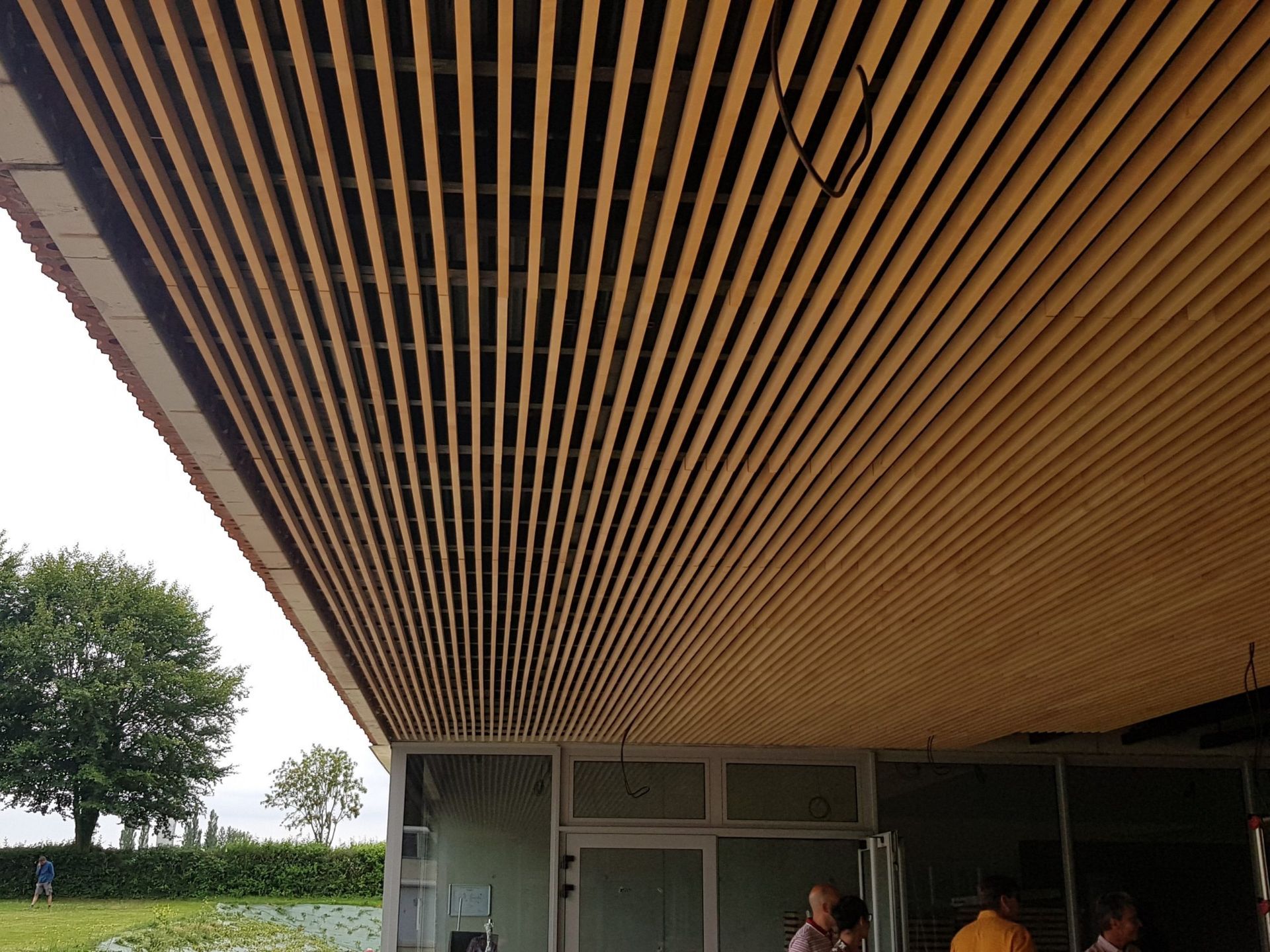 Plafond à lattes de bois surplombant l'extérieur d'un bâtiment. On voit des personnes à travers des portes vitrées.