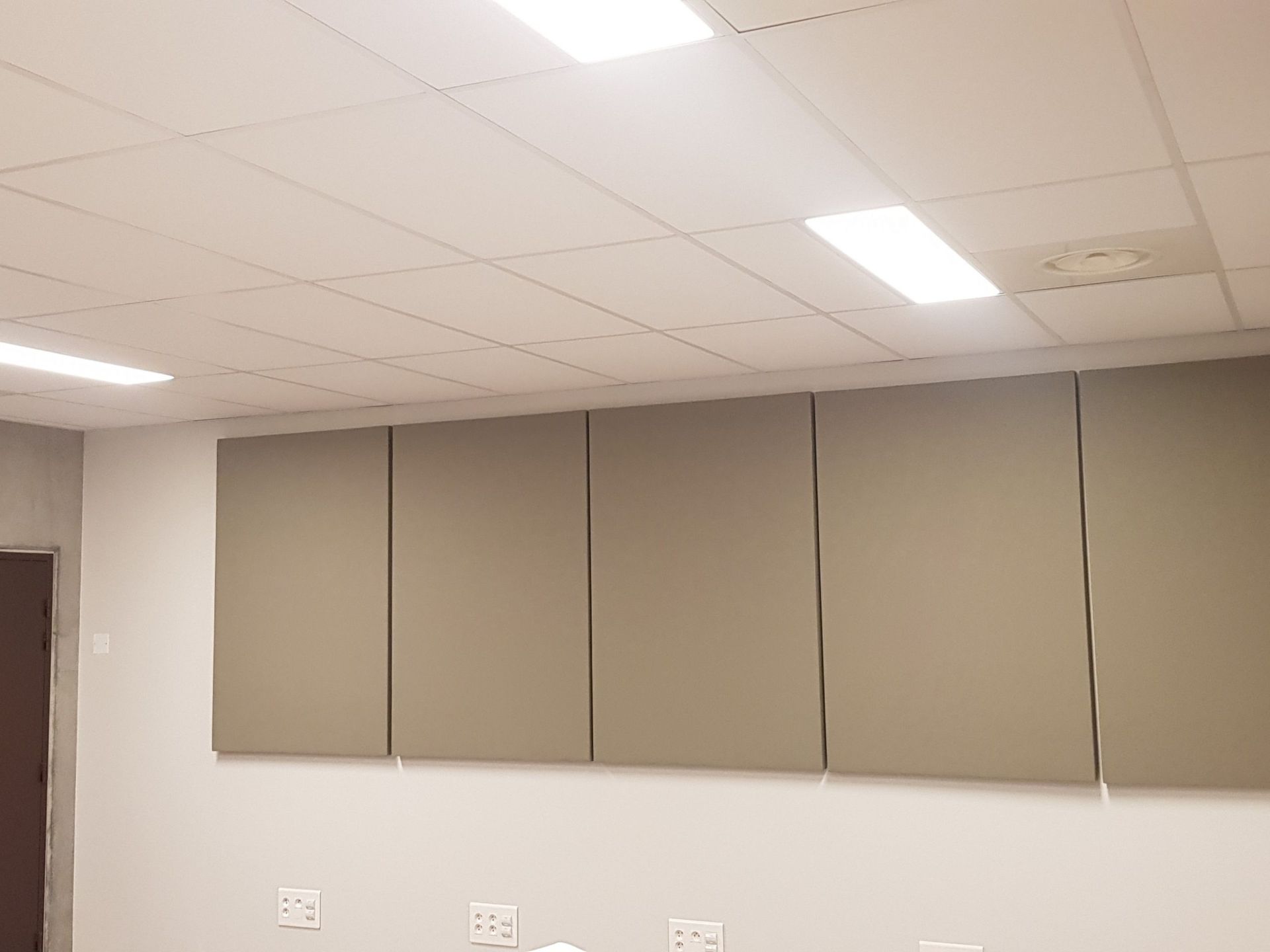Cinq panneaux d'insonorisation gris sur un mur blanc dans une pièce avec un plafond blanc et des plafonniers.