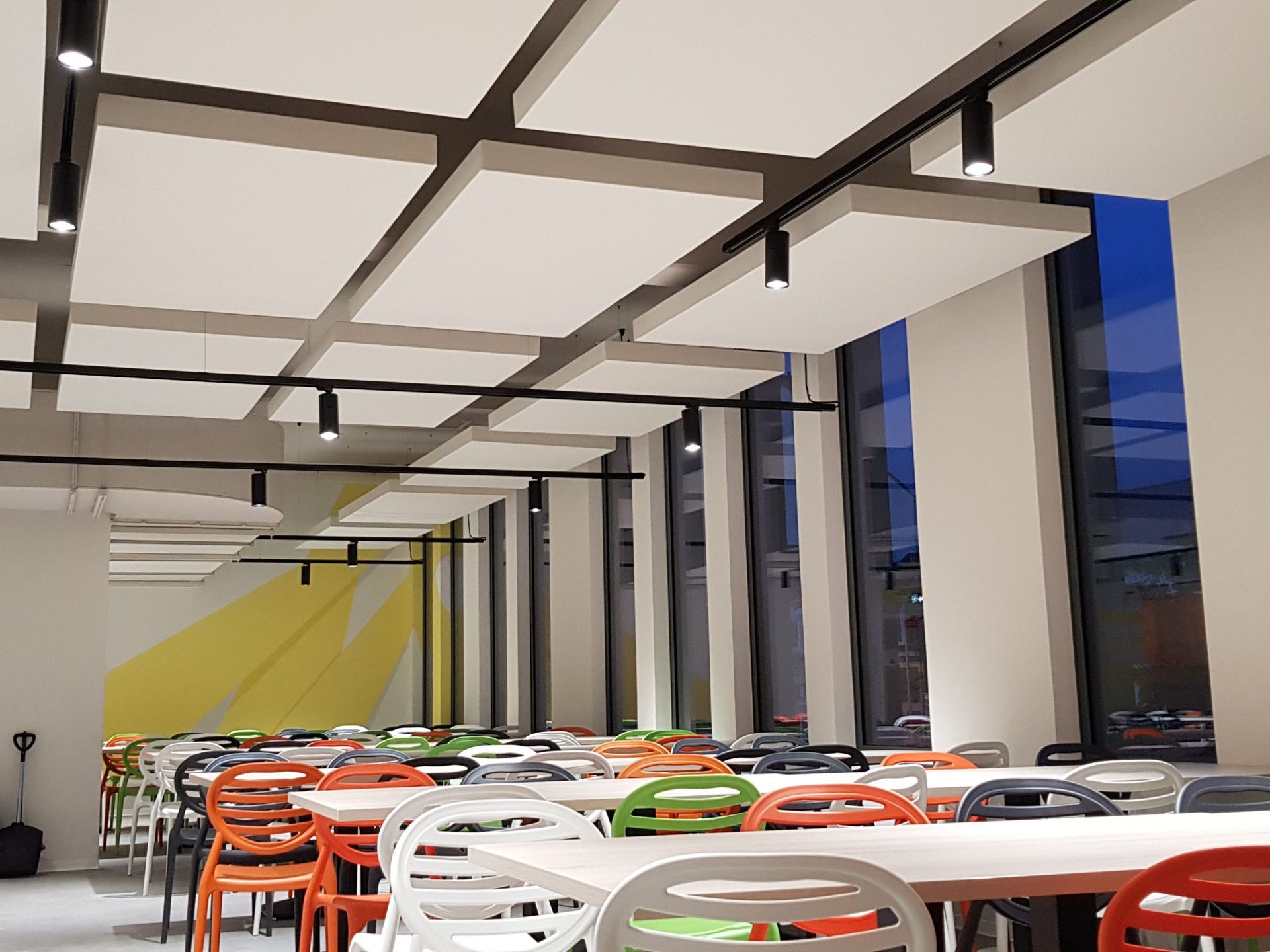 Cafétéria avec tables et chaises blanches, dalles de plafond acoustiques et grandes fenêtres.