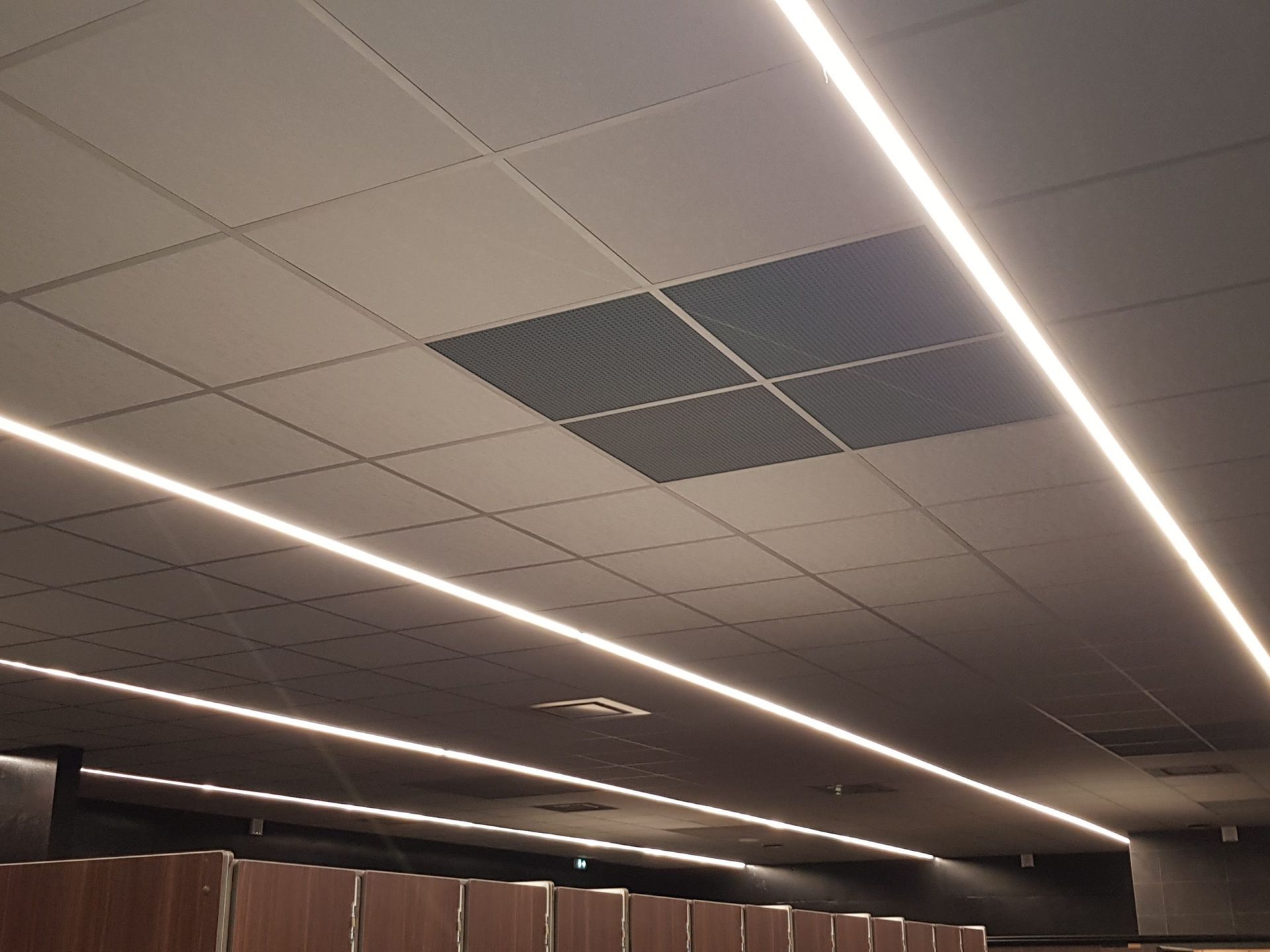 Plafond avec dalles carrées, lumières linéaires et panneau de ventilation carré foncé.