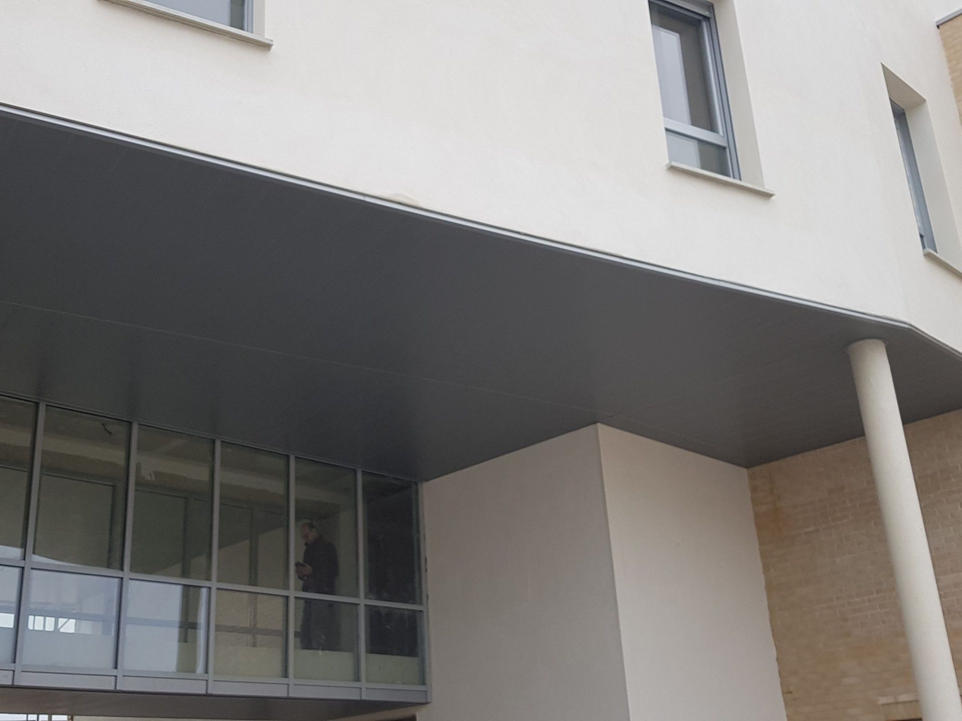 Extérieur d'un bâtiment moderne avec surplomb gris, fenêtres en verre et murs blancs et beiges.